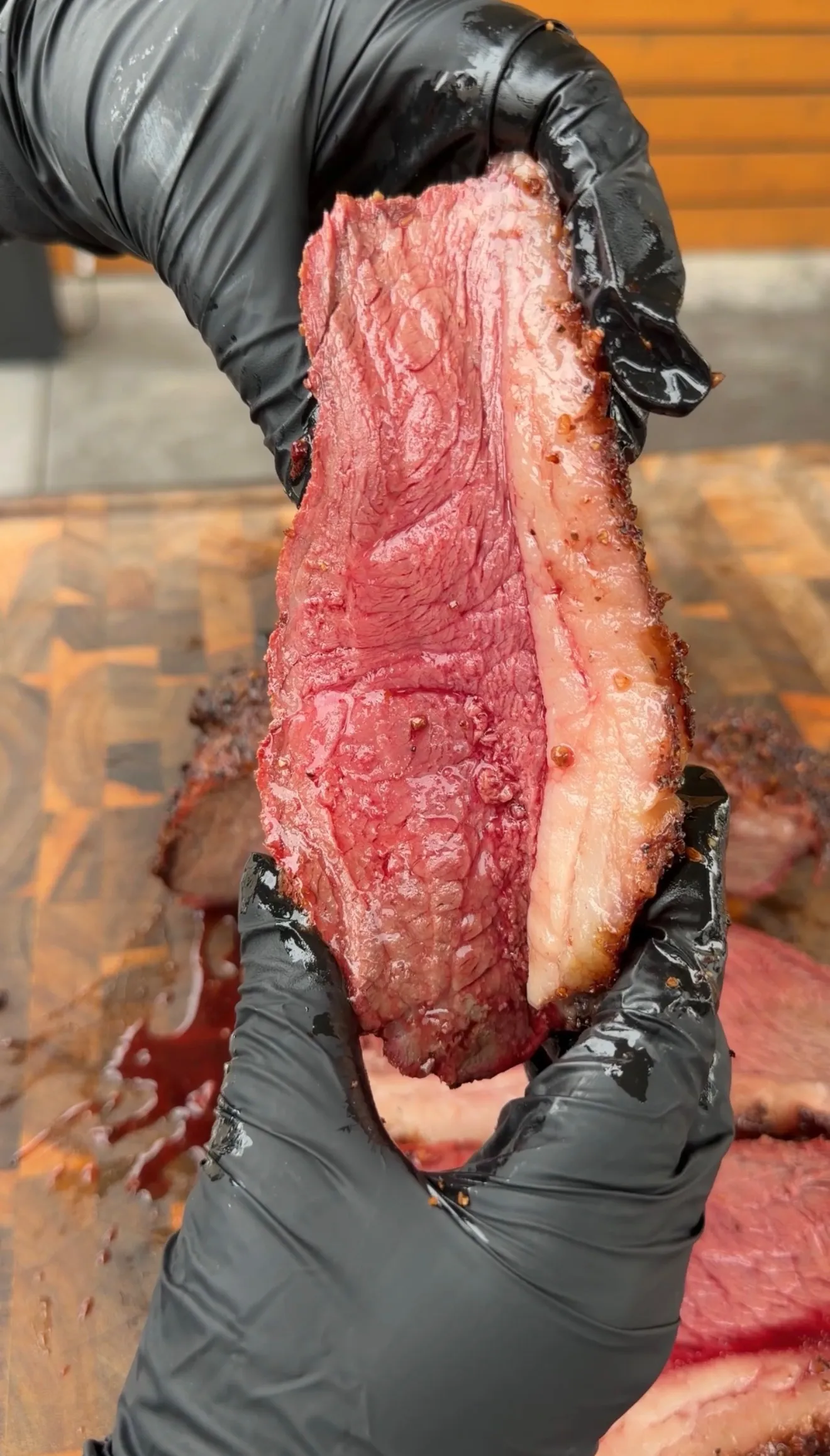 Gloved hands holding a thick slice of smoked picanha roast revealing a deep pink smoke ring and fat cap