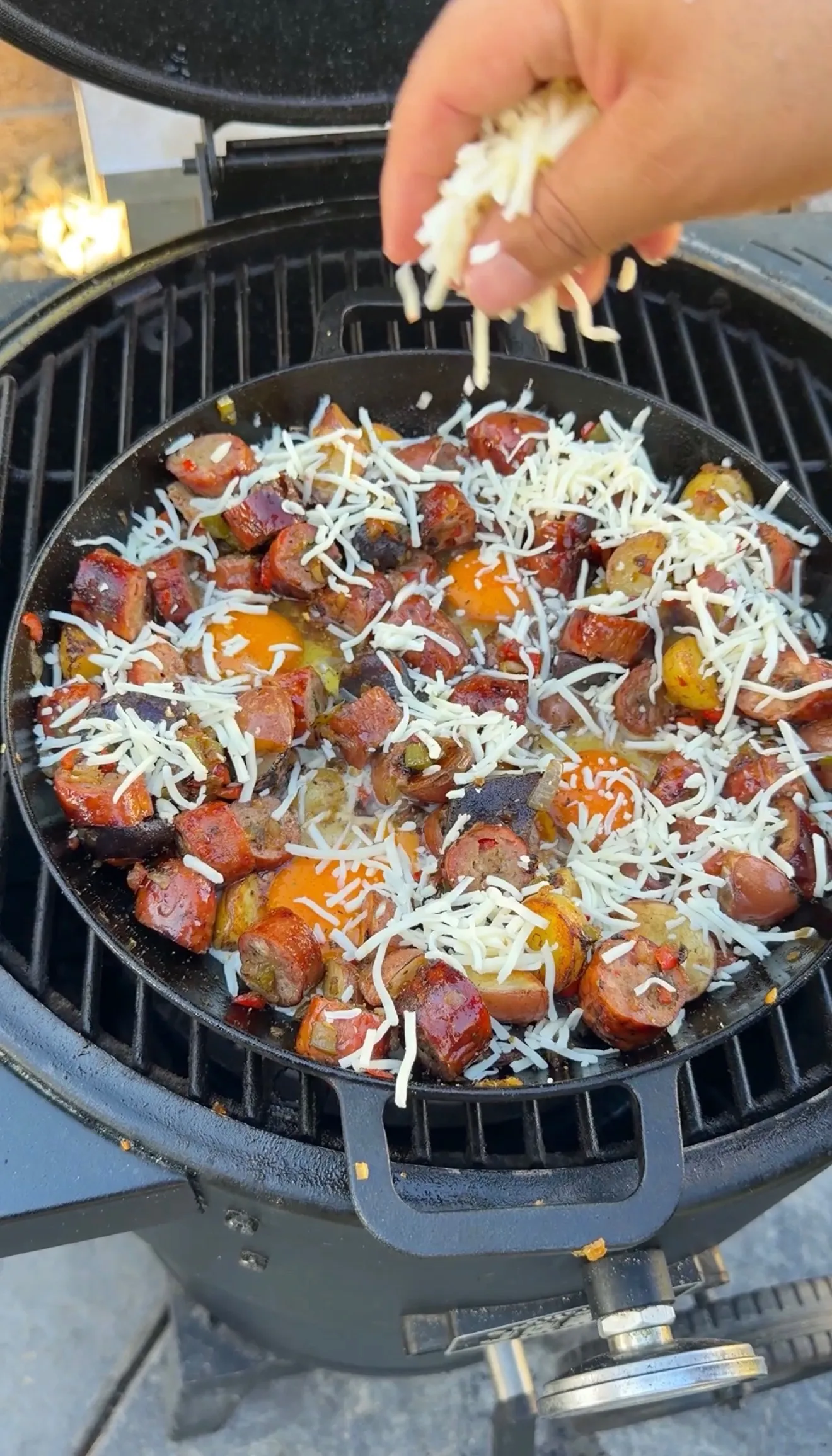 Hand sprinkling shredded mozzarella over smoked sausage breakfast skillet with eggs on a grill