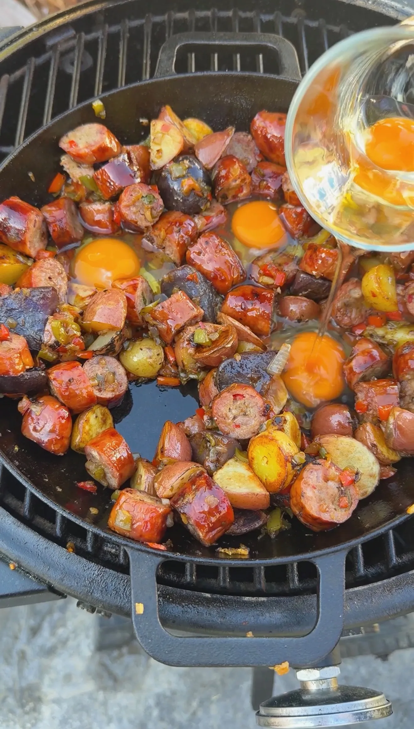 Eggs being poured into a cast iron skillet with smoked sausage, potatoes, and peppers on a grill