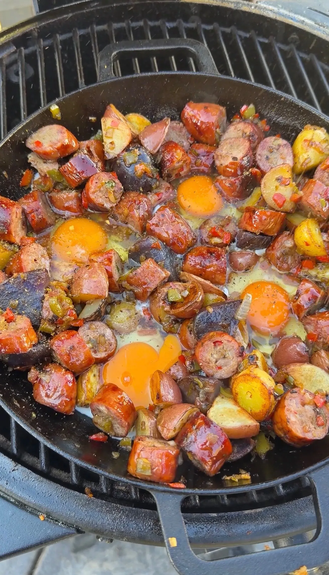 Cast iron skillet with smoked sausage, potatoes, peppers, and cracked eggs cooking on an outdoor grill