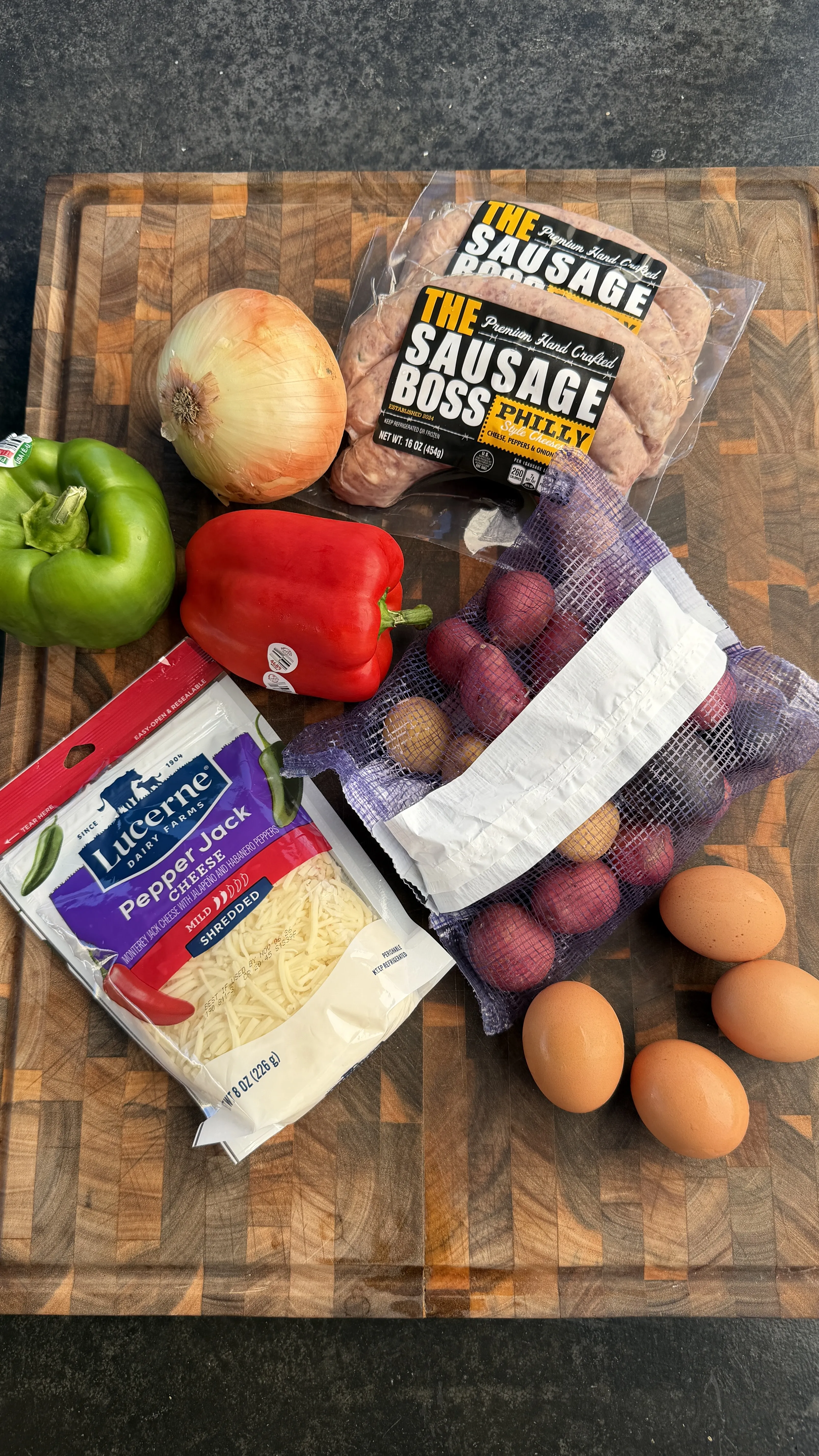 Breakfast skillet ingredients on a wooden board: Philly sausage, potatoes, eggs, bell peppers, onion, and shredded cheese