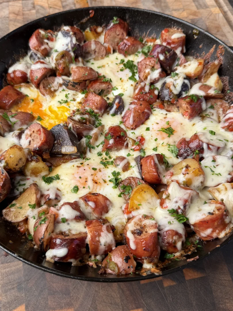 Cast iron skillet with smoked sausage, roasted potatoes, eggs, melted cheese, and fresh parsley garnish
