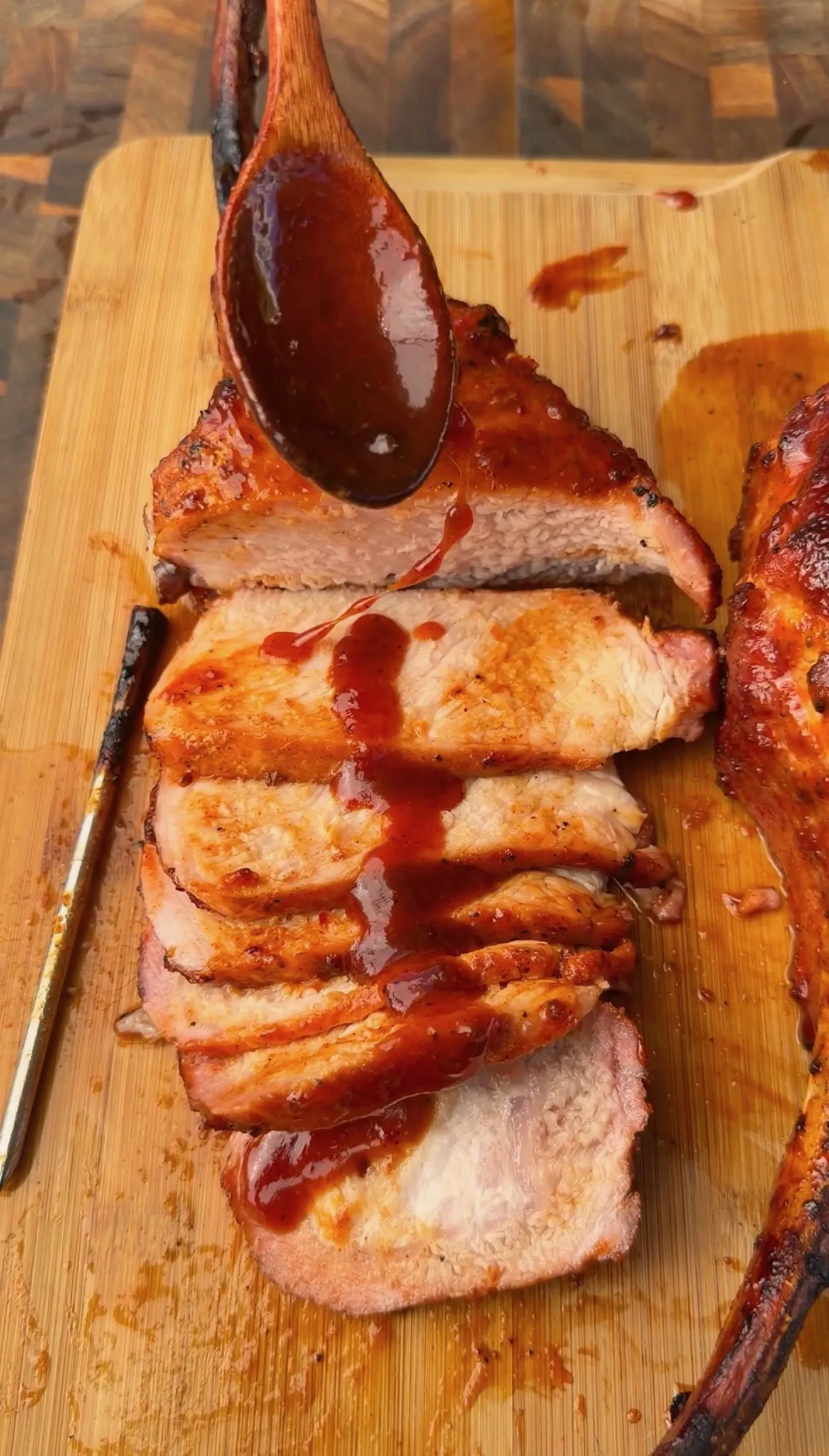 Sliced smoked tomahawk pork chop on wooden board drizzled with black raspberry BBQ sauce from a wooden spoon