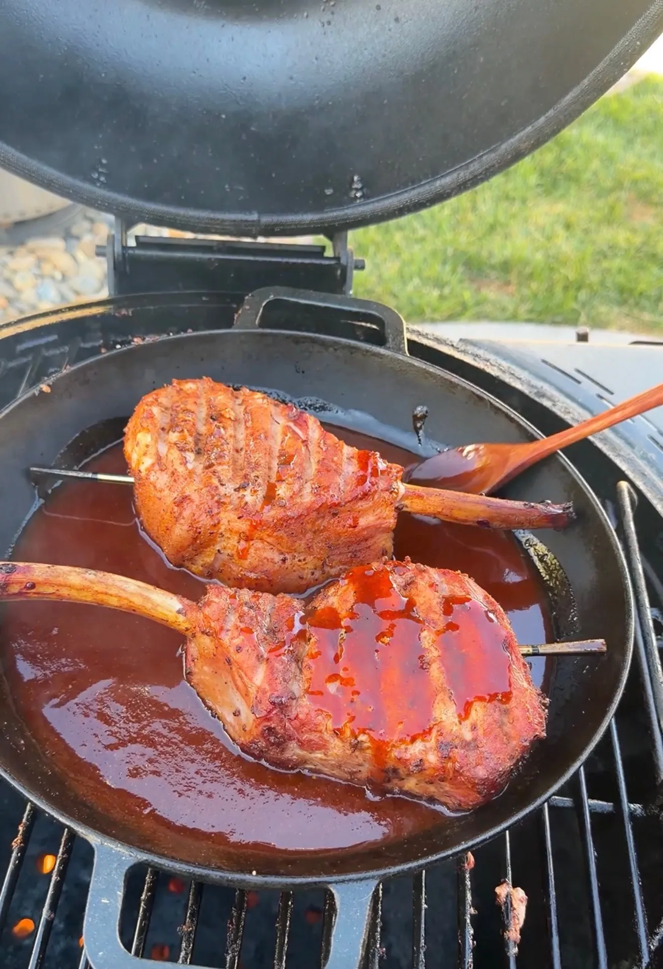 Two smoked tomahawk pork chops glazed in black raspberry BBQ sauce cooking in a cast iron skillet on a grill