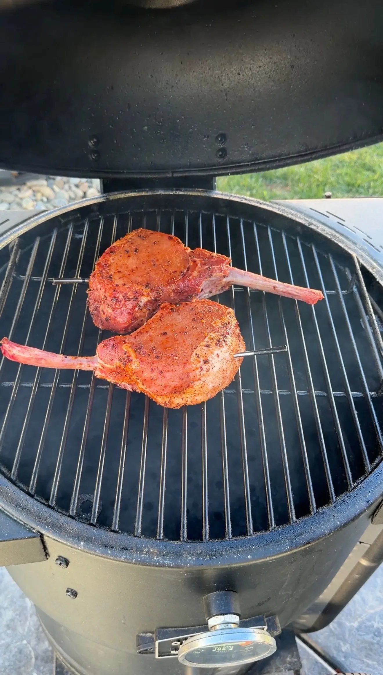 Two seasoned tomahawk pork chops with long bones smoking on a round ceramic kamado-style grill outdoors