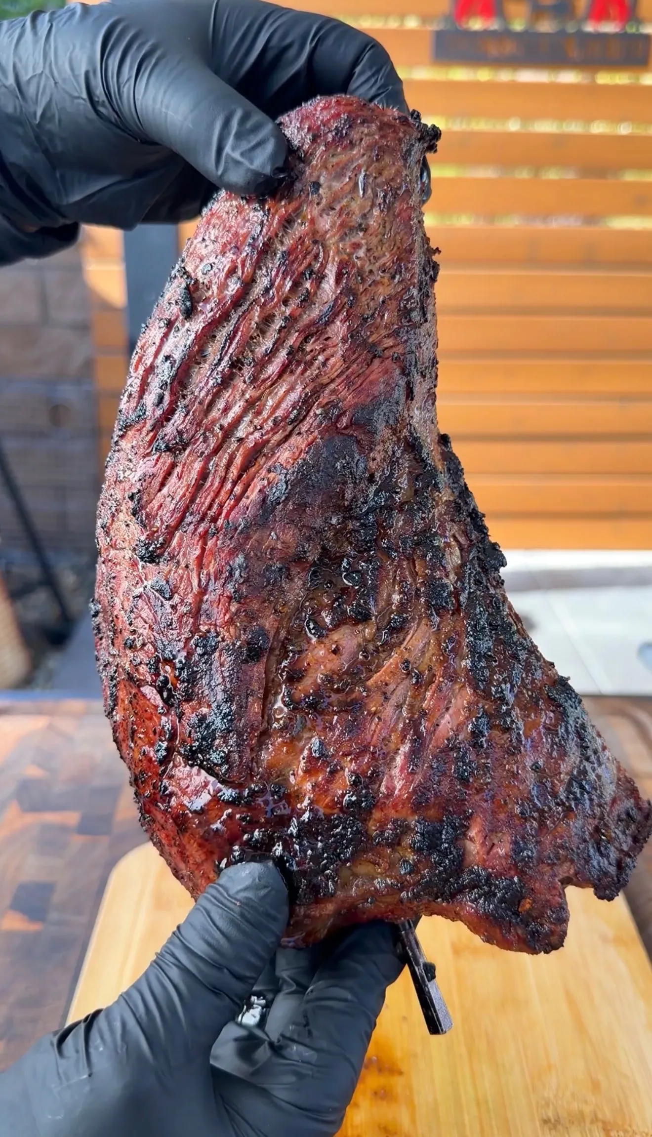 Hands in black gloves holding a large smoked tri-tip with a dark, caramelized bark crust over a wooden cutting board