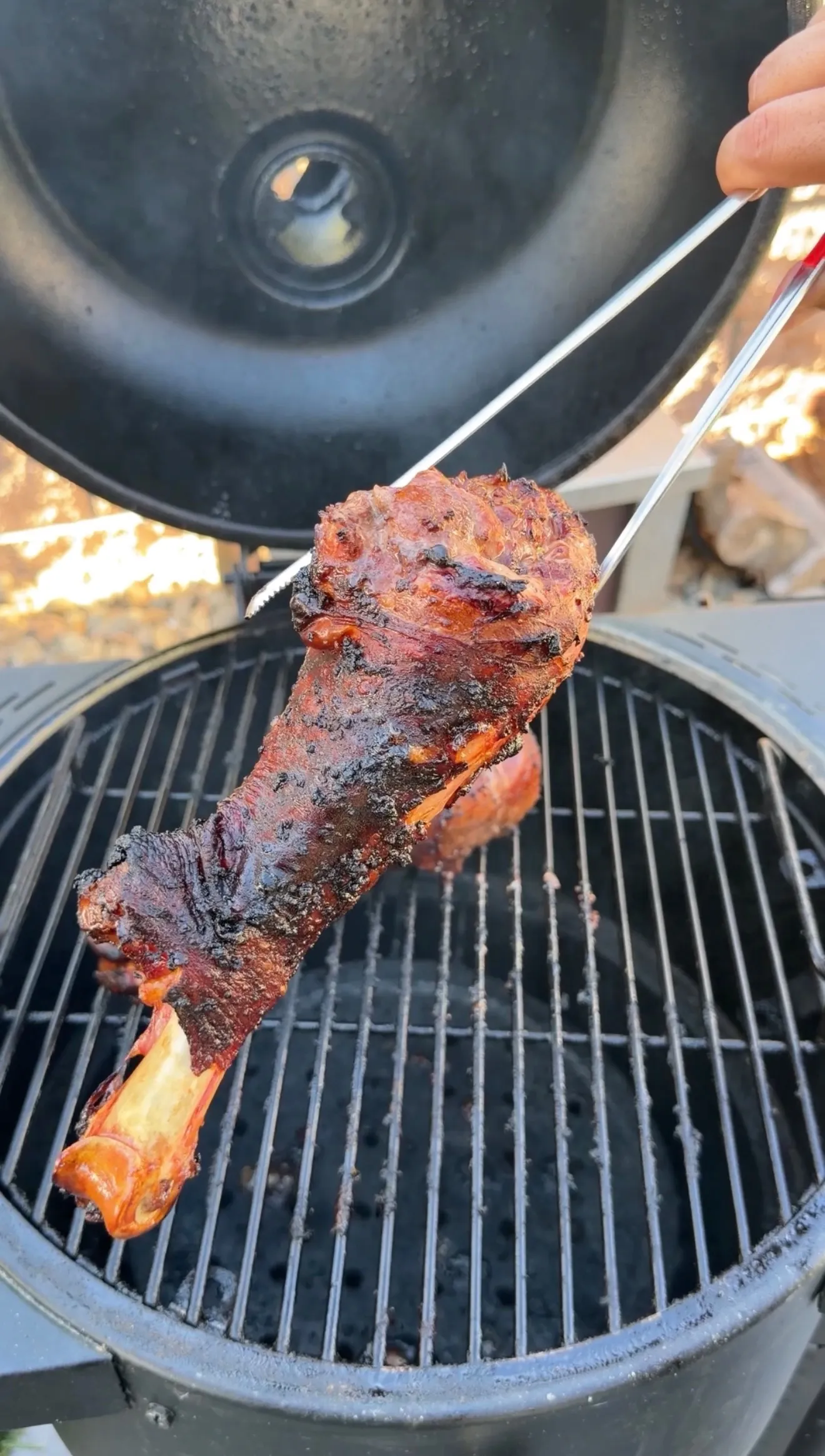 A fall-apart smoked turkey leg with dark bark being lifted by tongs over a round charcoal grill