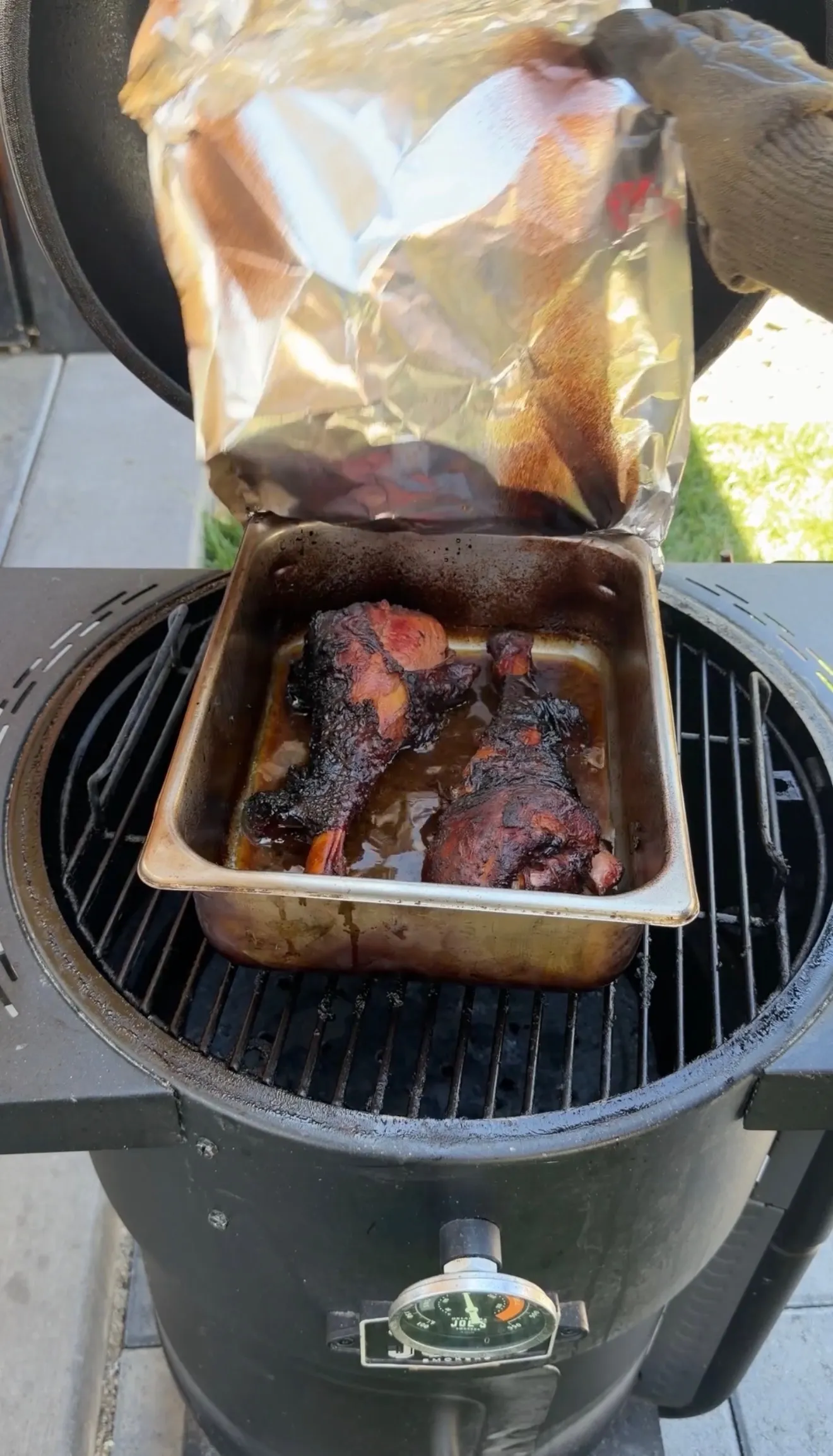 Fall-apart smoked turkey legs in a foil pan on a charcoal smoker, unwrapped from aluminum foil