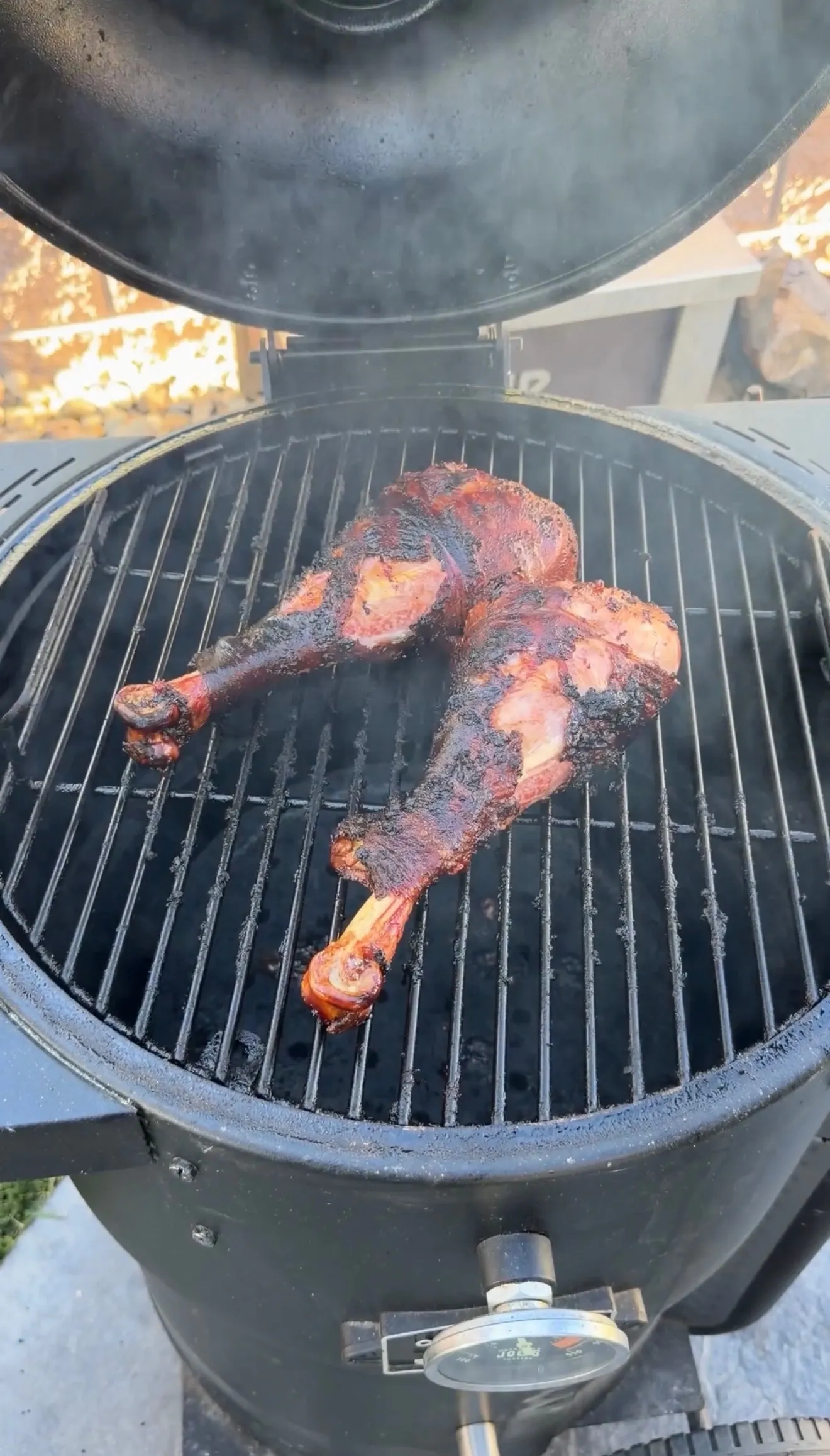 Two fall-apart smoked turkey legs with dark bark resting on a round charcoal smoker grill with smoke rising