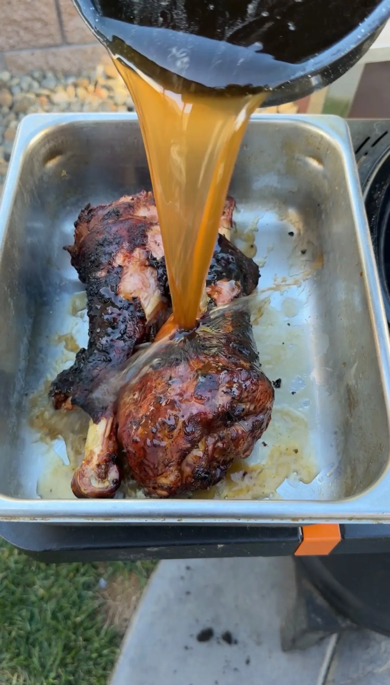 Rich amber BBQ sauce being poured over fall-apart smoked turkey legs in a stainless steel roasting pan