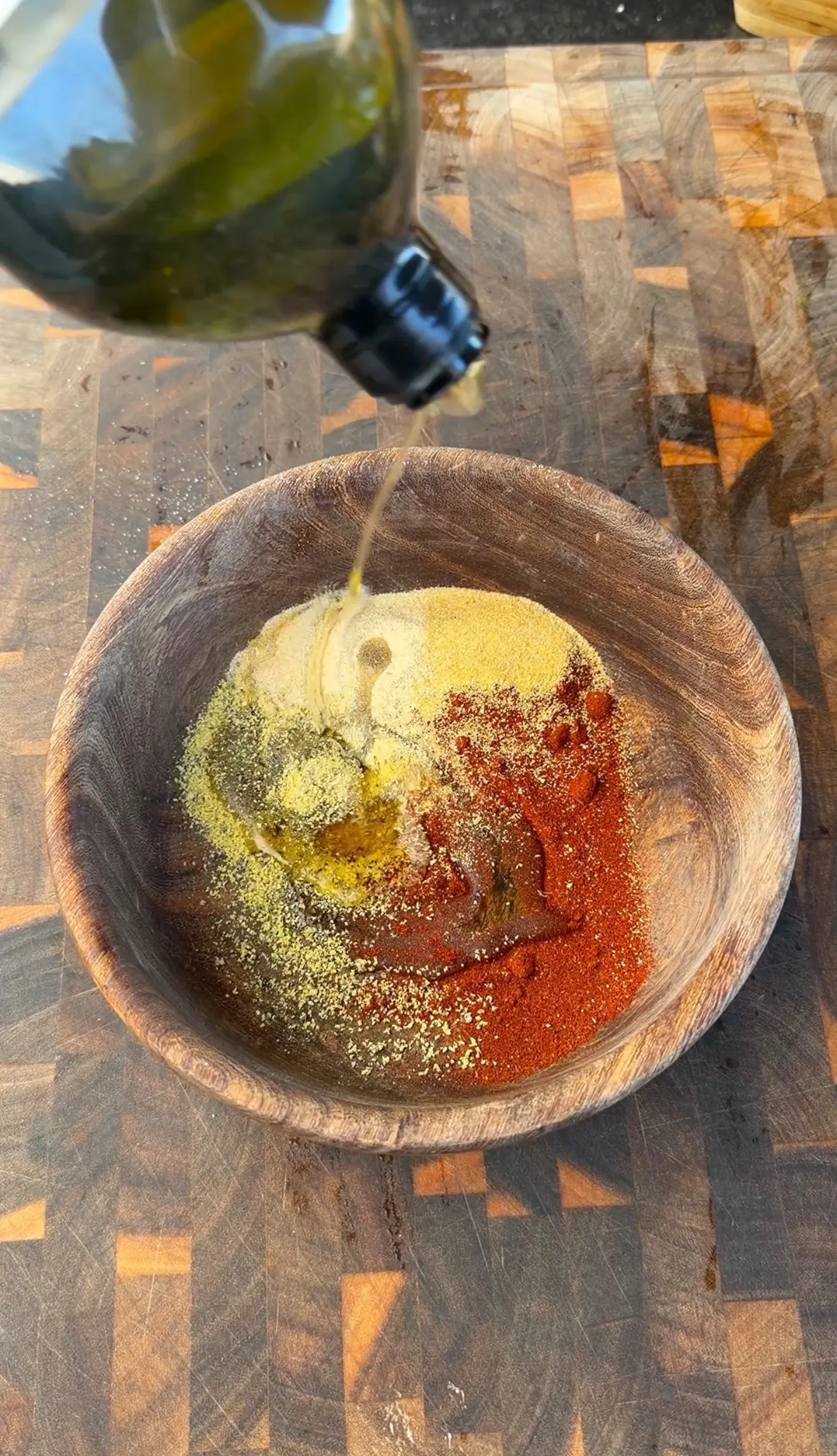 Olive oil being poured over paprika, garlic powder, and lemon pepper spices in a rustic wooden bowl