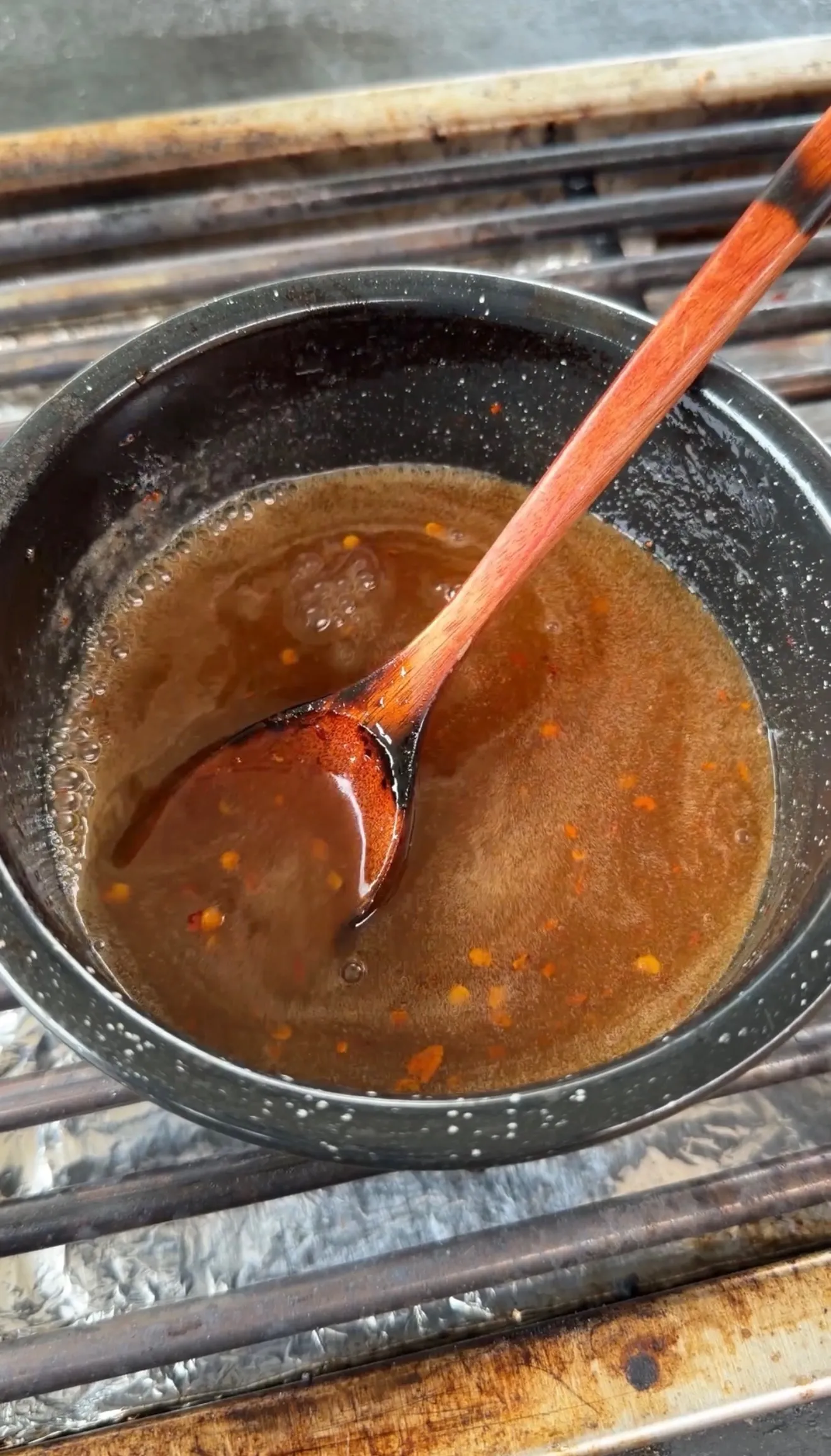 Bubbling spicy hot sauce glaze with red pepper flakes in a black saucepan with a wooden spoon on a grill