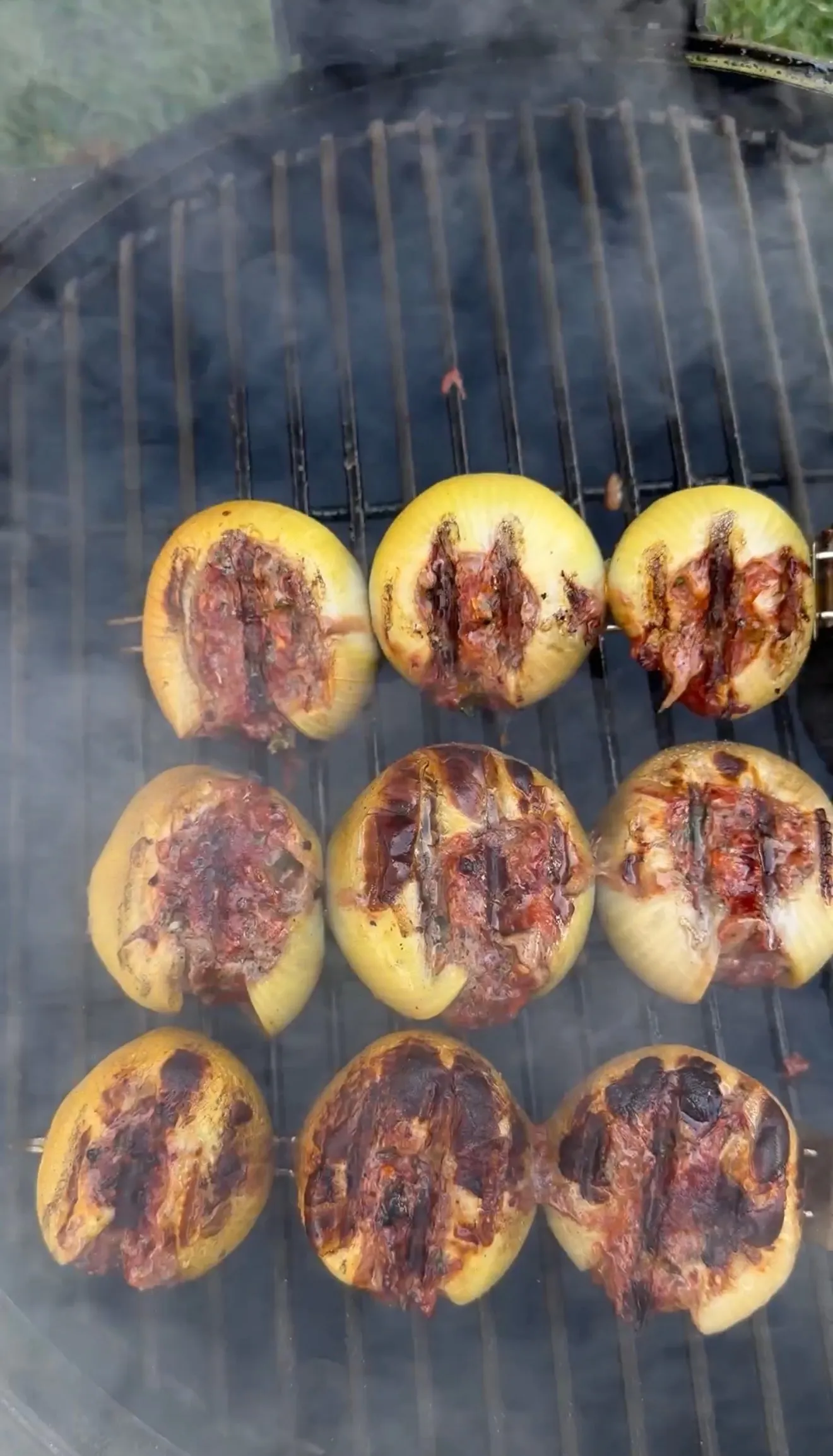 Nine meat-stuffed onion bombs with grill marks smoking on a charcoal grill grate