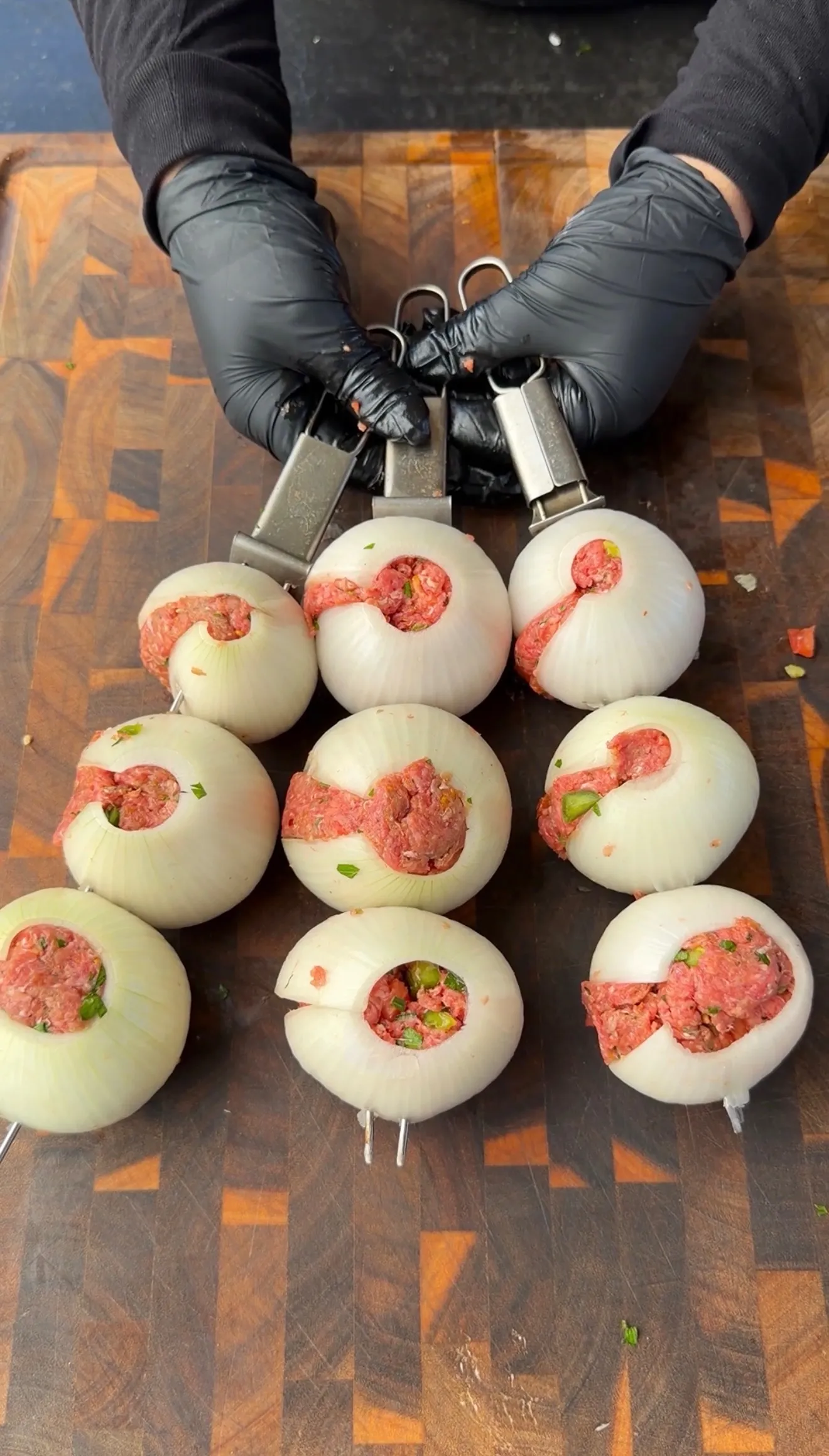 Whole white onions stuffed with seasoned ground meat mixture on metal skewers on a wooden cutting board
