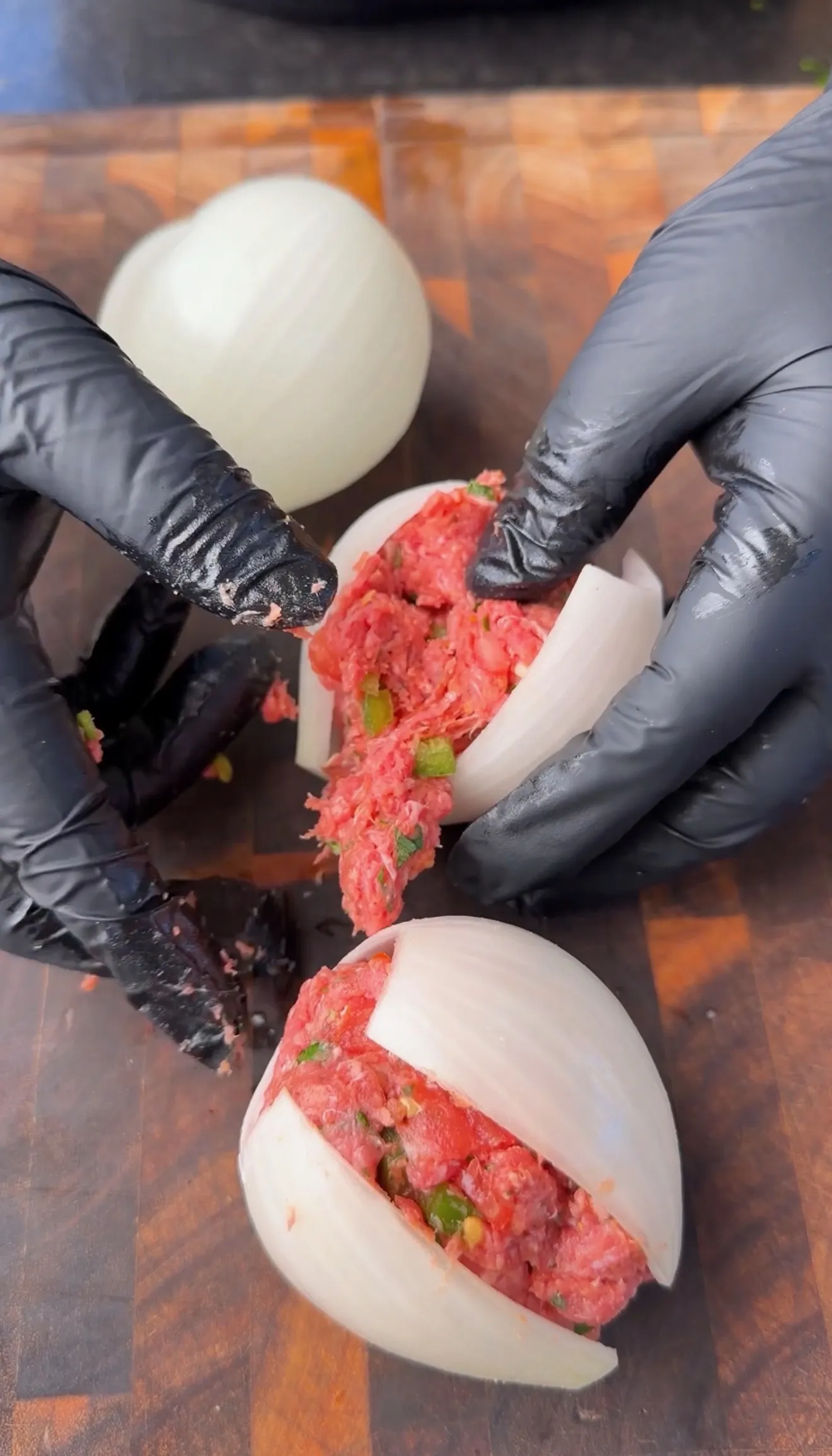 Hands in black gloves stuffing hollowed white onions with seasoned raw ground beef mixture on a wooden cutting board
