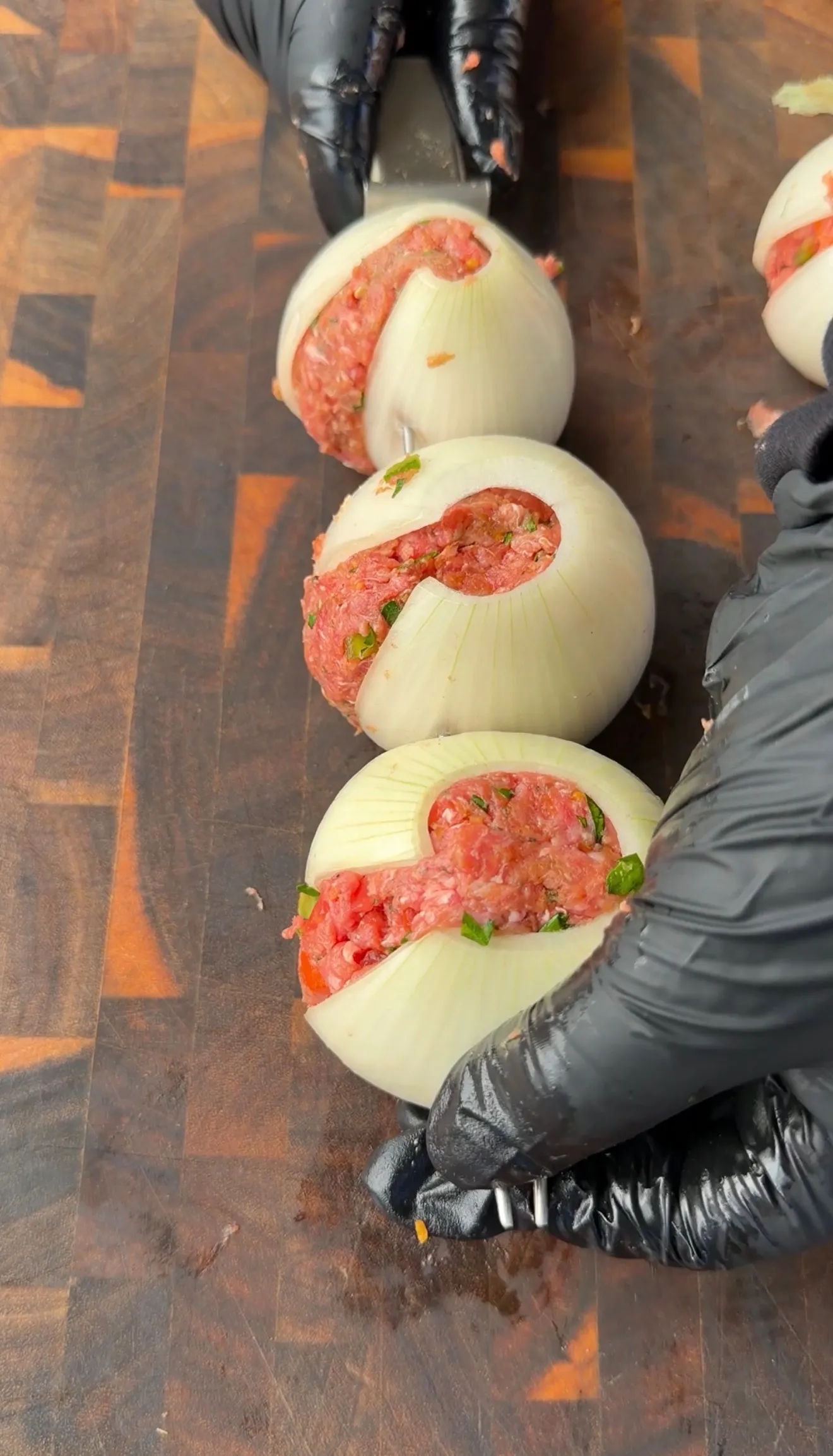 Whole white onions stuffed with seasoned ground meat mixture on a wooden cutting board, held by gloved hands