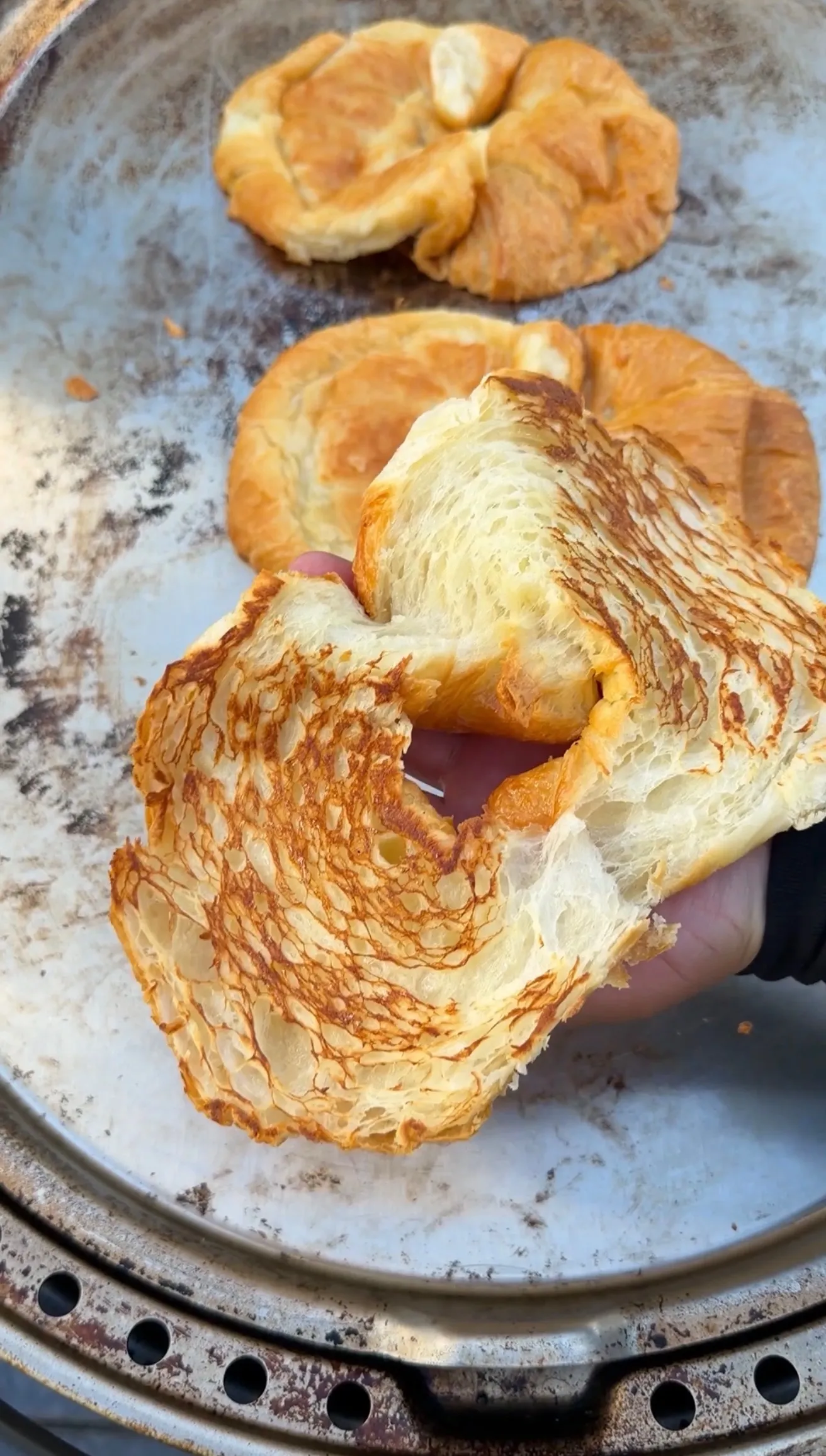 Hand holding a toasted croissant sliced open as a burger bun, with more croissants on a baking tray