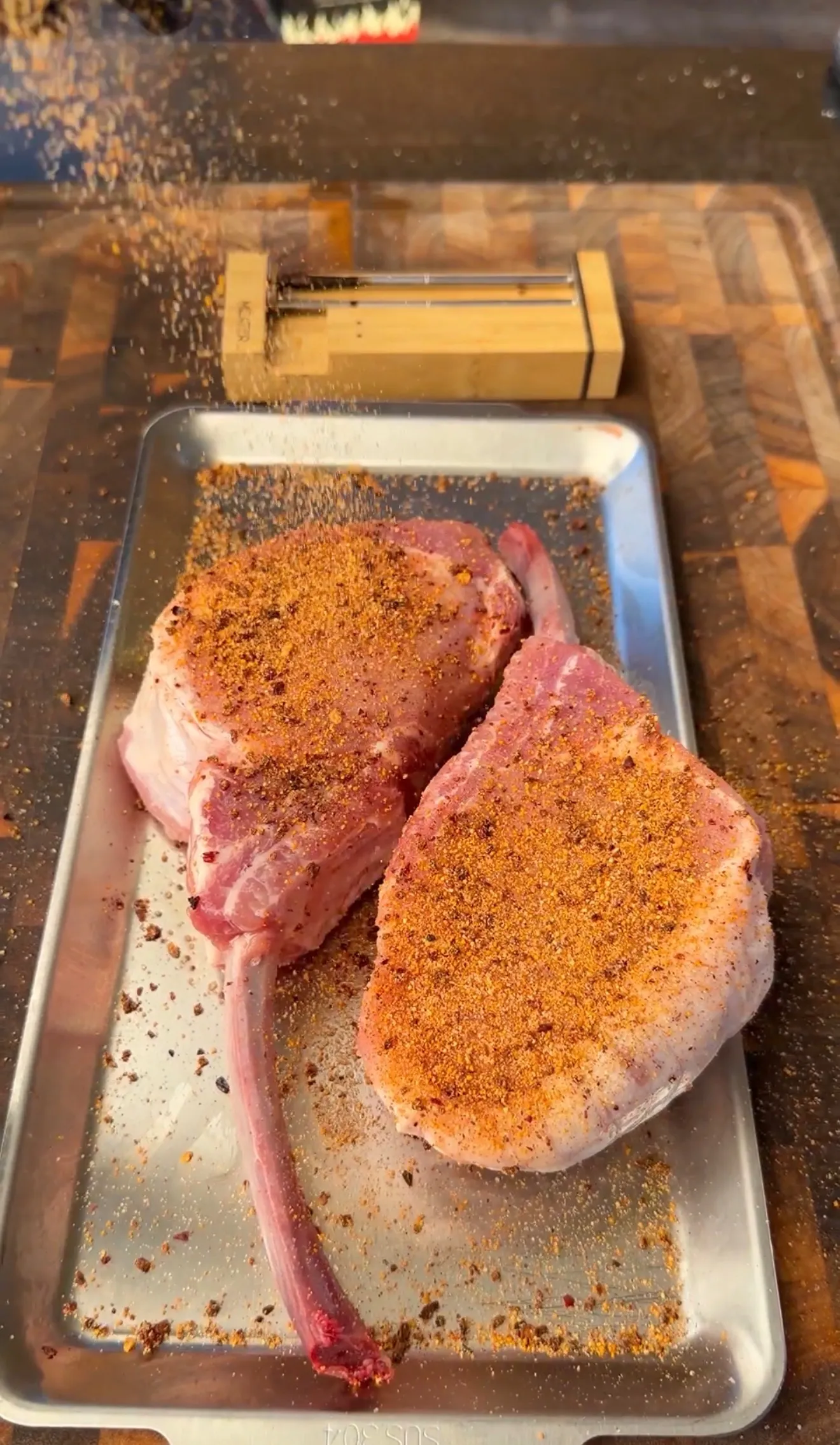 Two raw tomahawk pork chops coated in orange spice dry rub on a metal baking sheet, ready for smoking