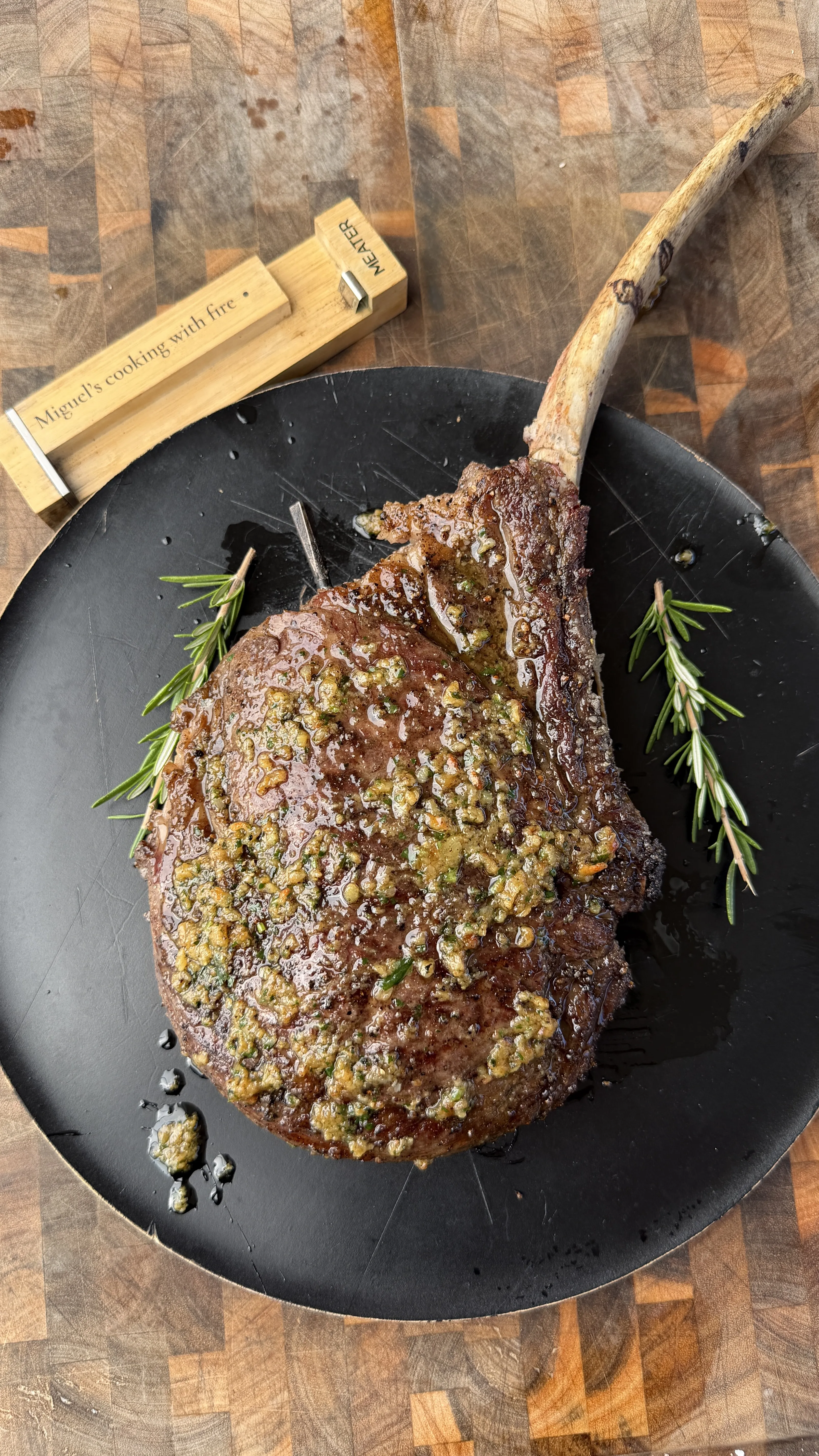 Juicy tomahawk ribeye steak topped with herb garlic butter on a black plate with fresh rosemary sprigs