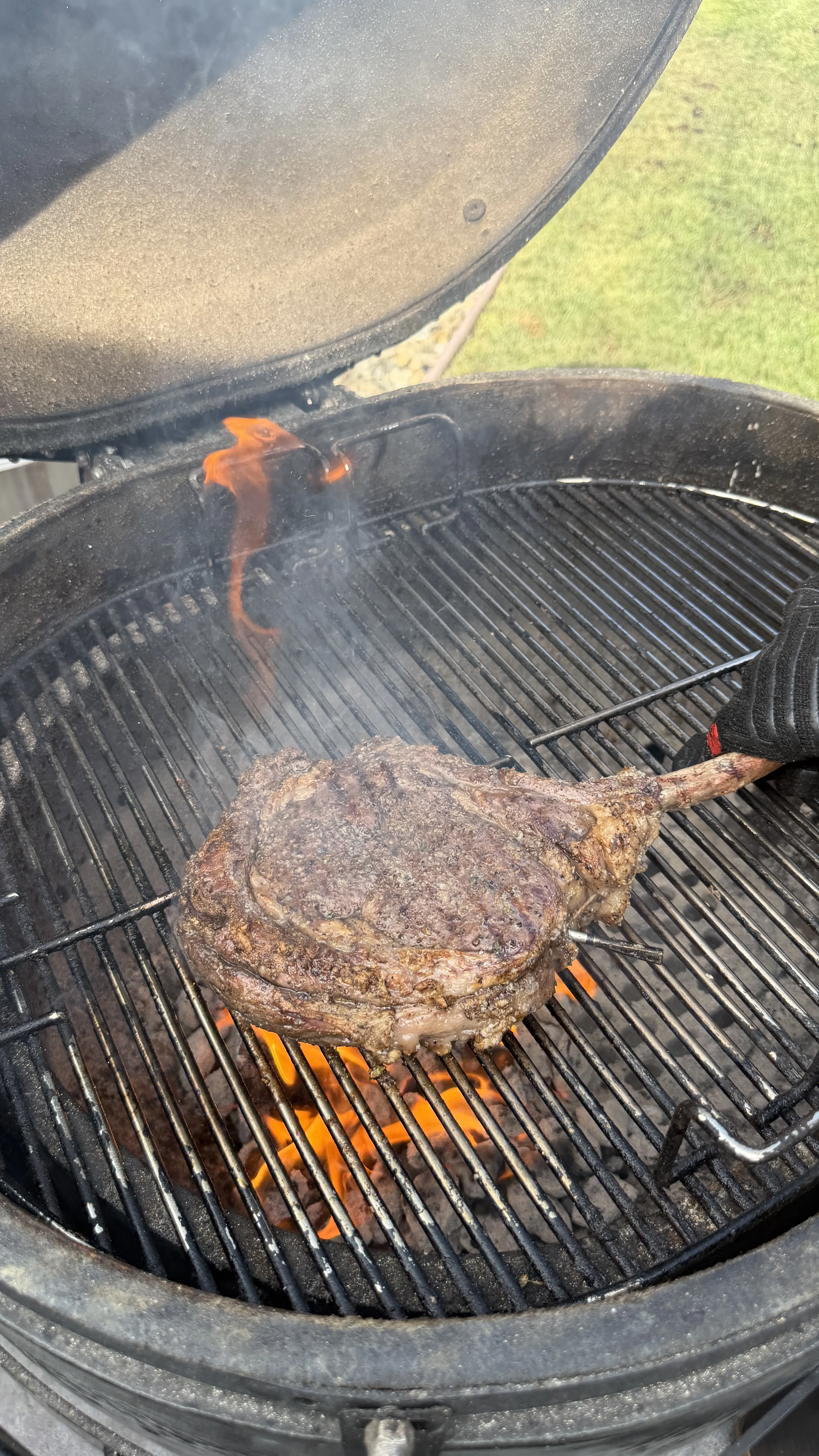 Thick tomahawk ribeye steak with a long bone searing over open flames on a charcoal kettle grill