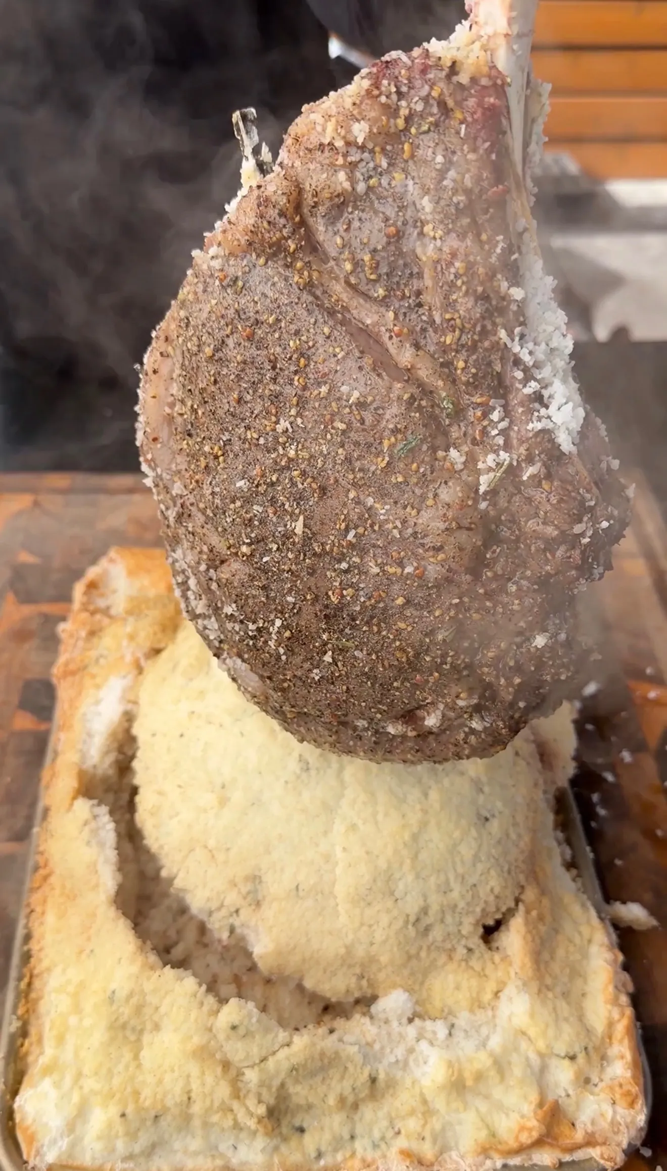 Seasoned tomahawk ribeye steak being lifted from a salt crust casing with smoke rising around it
