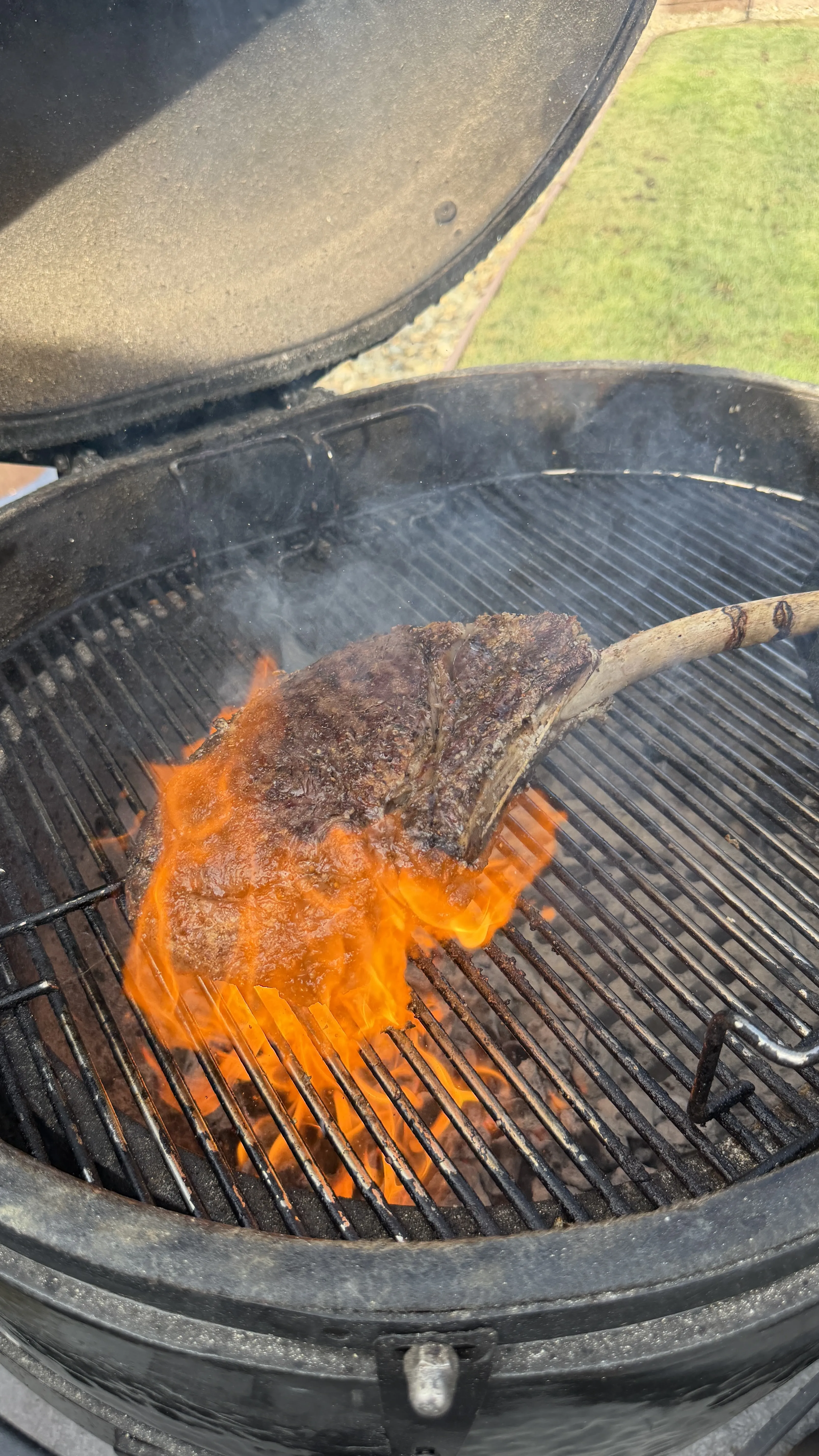 Thick tomahawk ribeye steak searing over open flames on a charcoal kamado grill with smoke rising