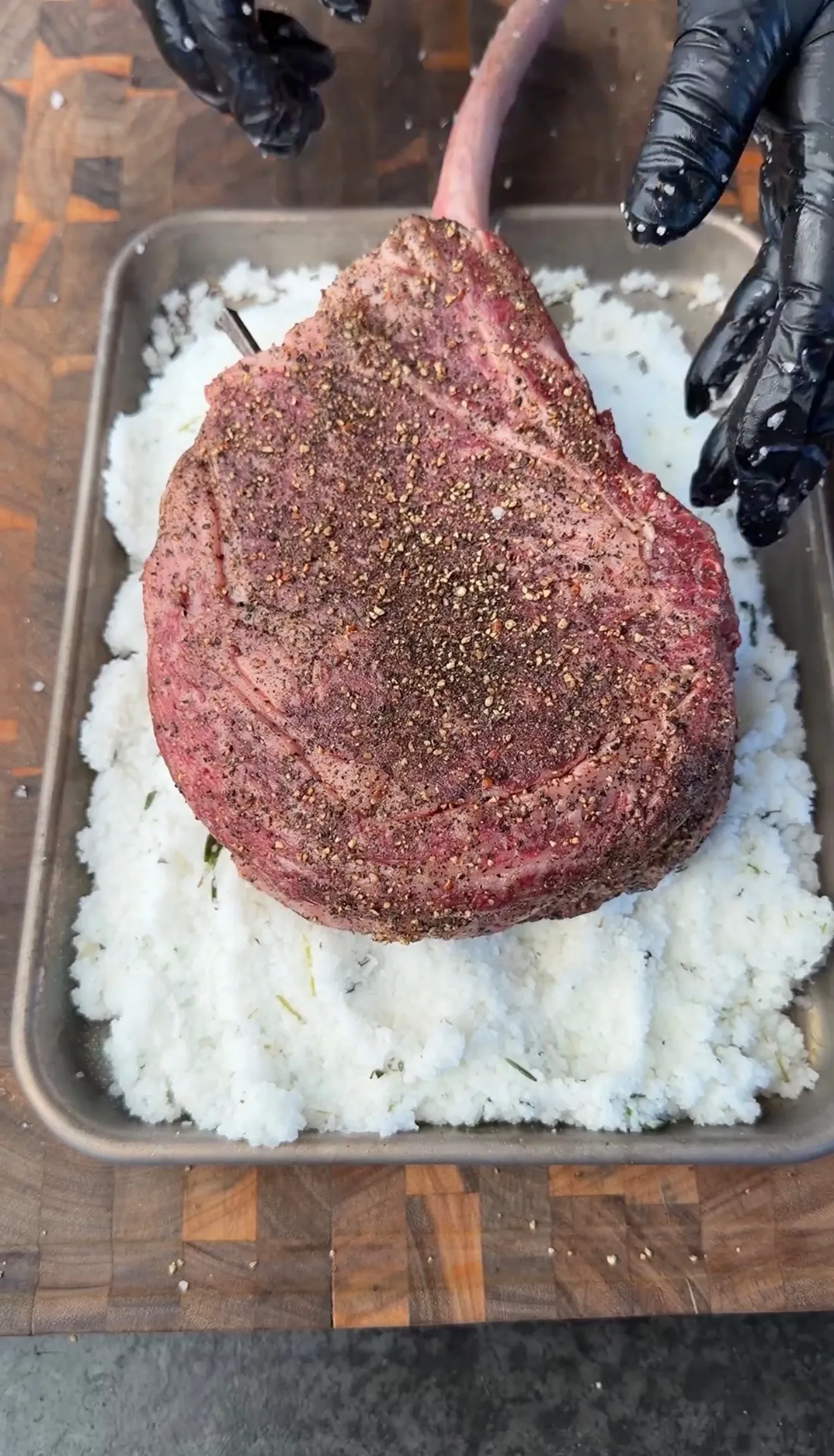 Large tomahawk ribeye steak coated in black pepper seasoning placed on a bed of coarse salt in a baking pan