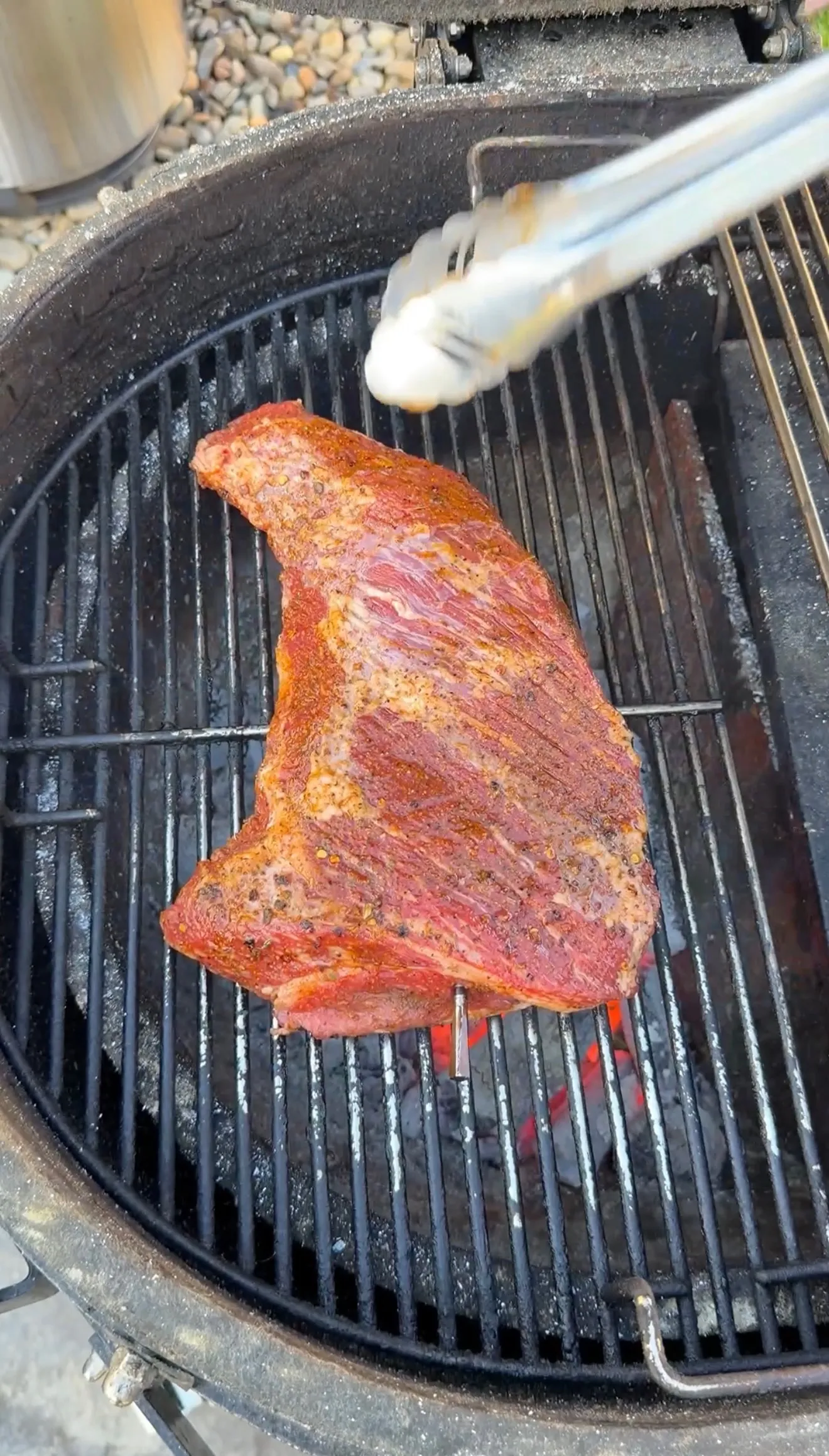 Seasoned tri-tip steak grilling over hot charcoal on a round kettle grill, held with metal tongs