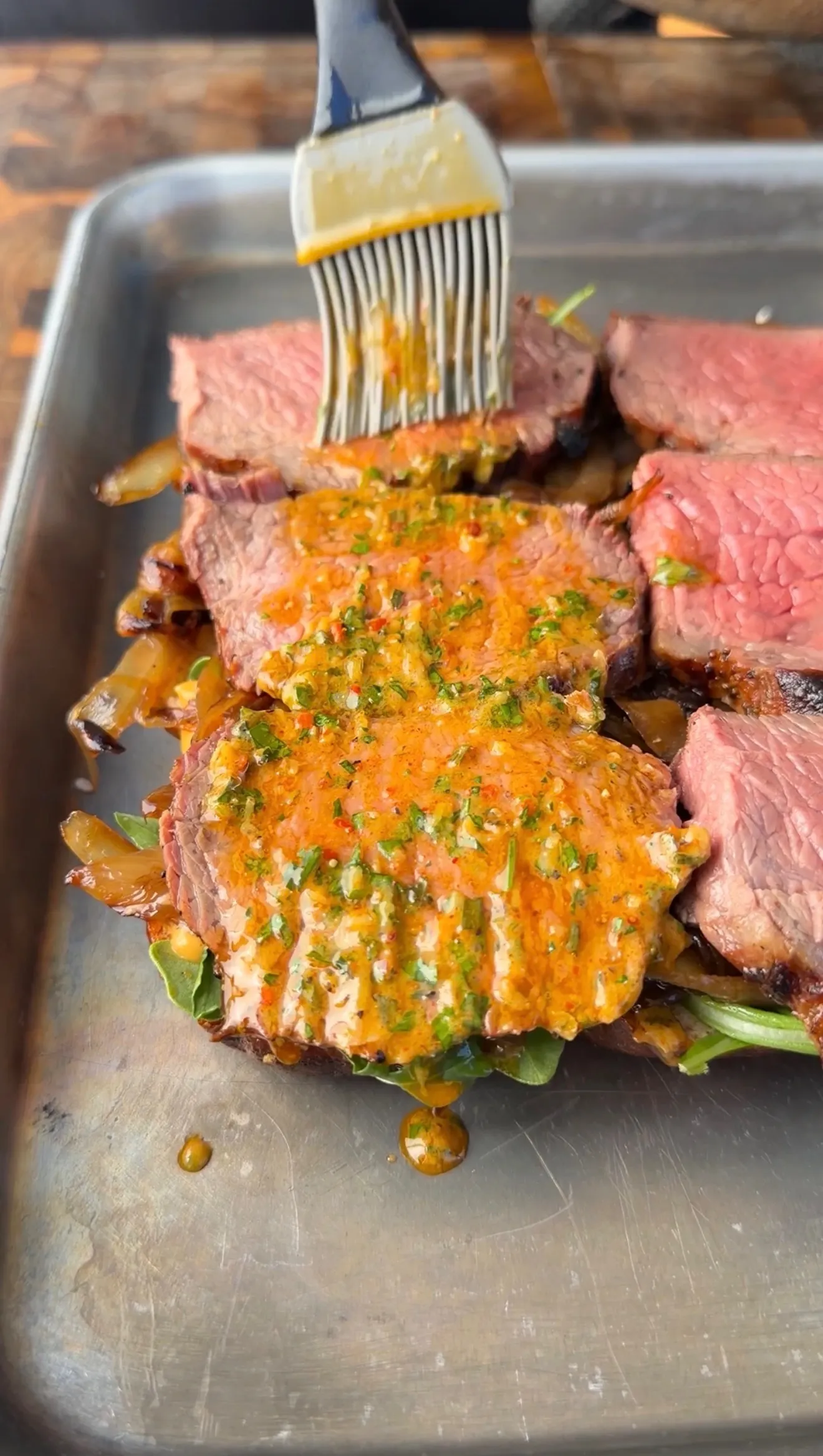Sliced tri-tip steak being brushed with vibrant orange cowboy butter sauce on a metal sheet pan