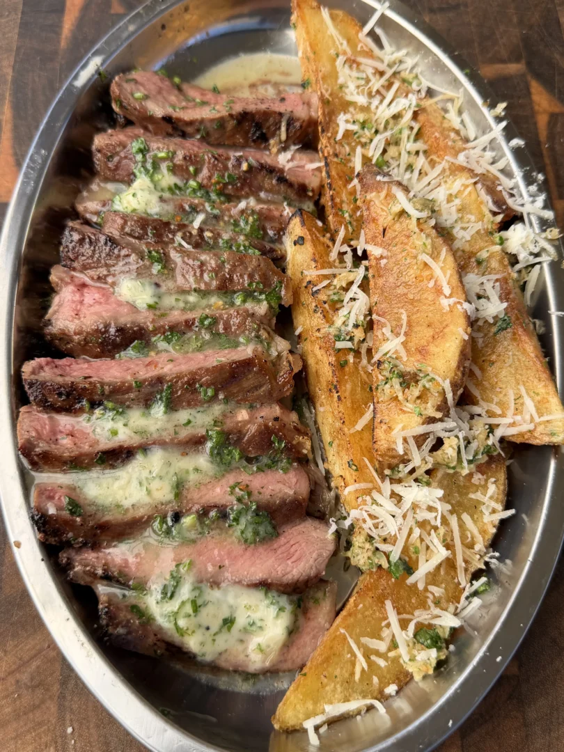 Sliced grilled ribeye steak with melted umami herb butter and crispy Parmesan potato wedges on a silver platter