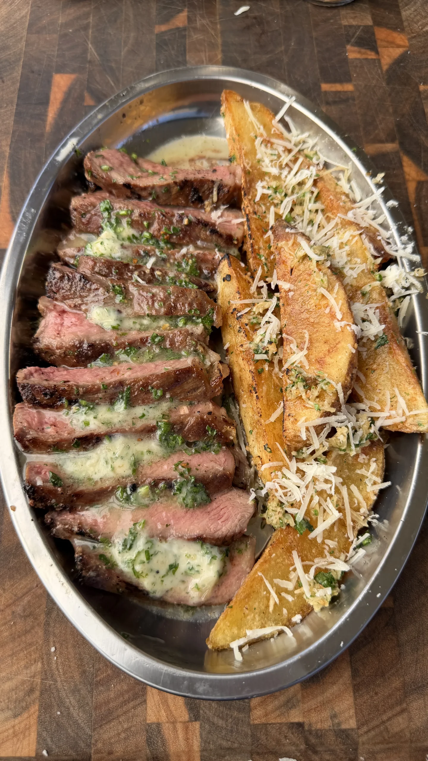 Sliced grilled ribeye steak with melted umami herb butter and crispy Parmesan potato wedges on a silver platter