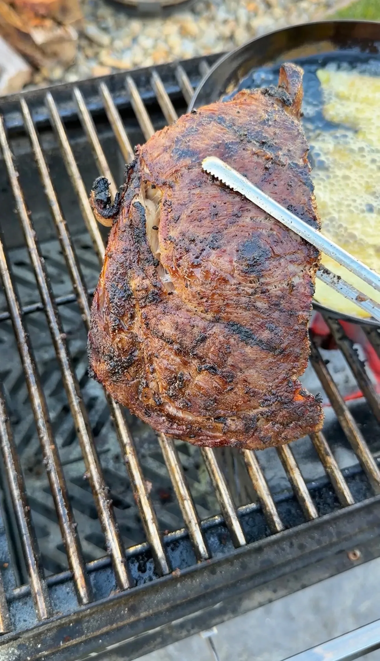 Thick ribeye steak with charred crust being grilled on outdoor barbecue grates with tongs