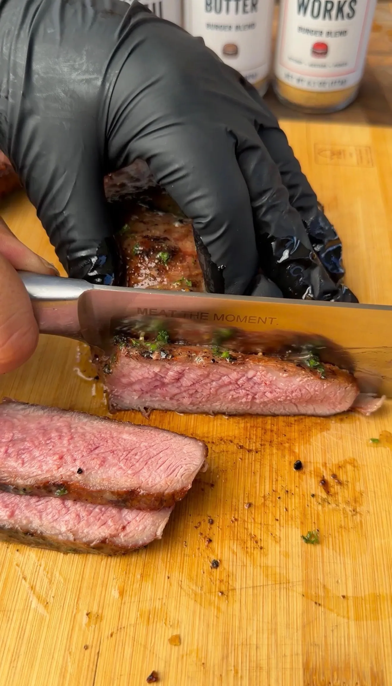 Gloved hands slicing a grilled ribeye steak to reveal a pink medium-rare center on a wooden cutting board
