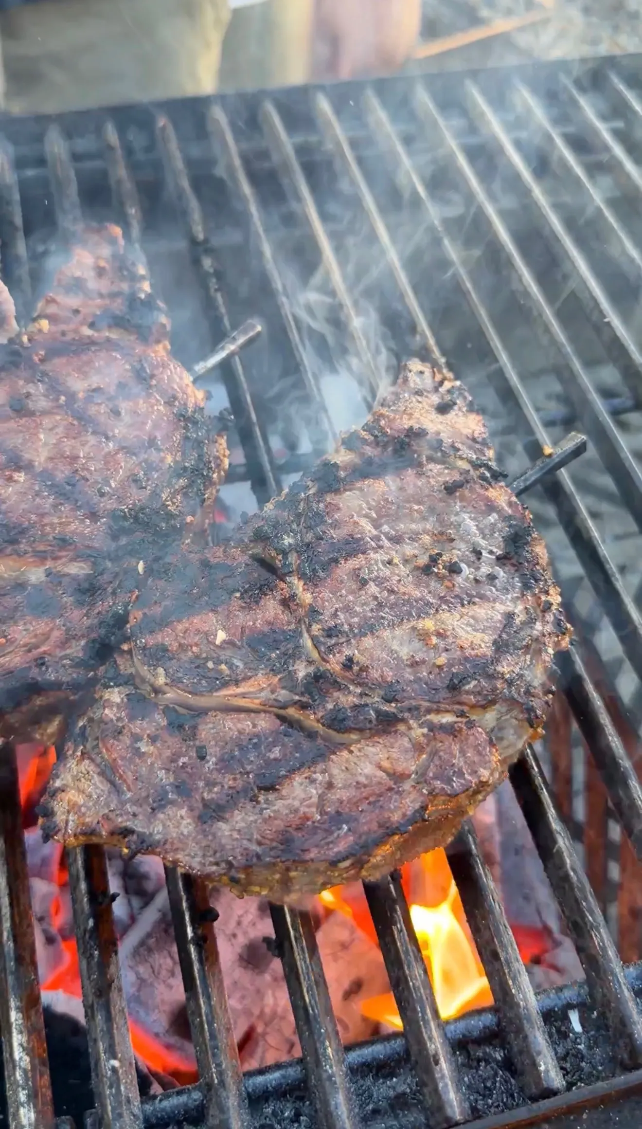 Thick ribeye steaks with charred grill marks smoking over glowing charcoal flames on an open grill
