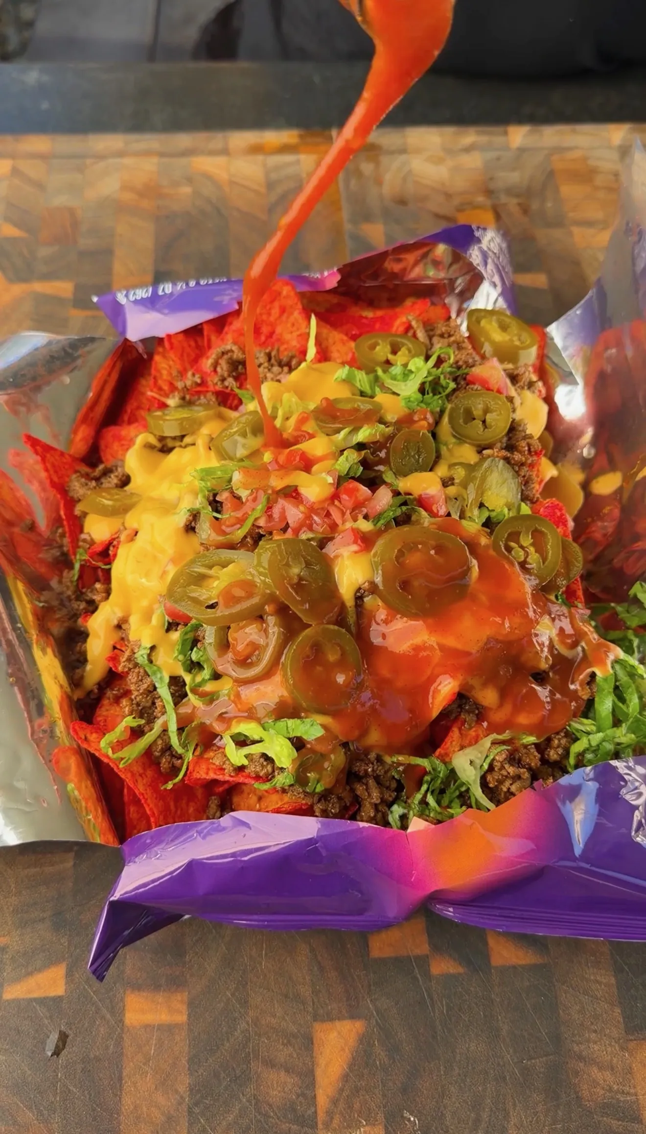 Walking taco in open Doritos bag topped with cheese sauce, jalapeños, ground beef, lettuce, and hot sauce being poured