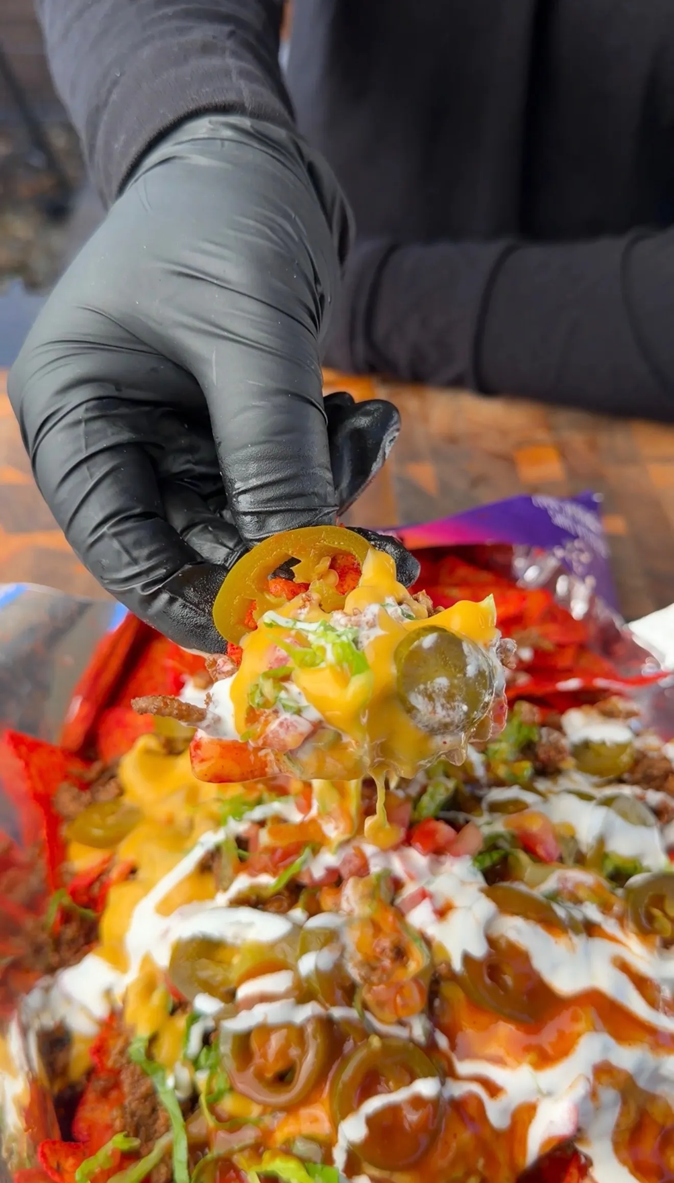 Gloved hand pulling a loaded chip from walking tacos topped with melted cheese, jalapeños, sour cream, and pico de gallo