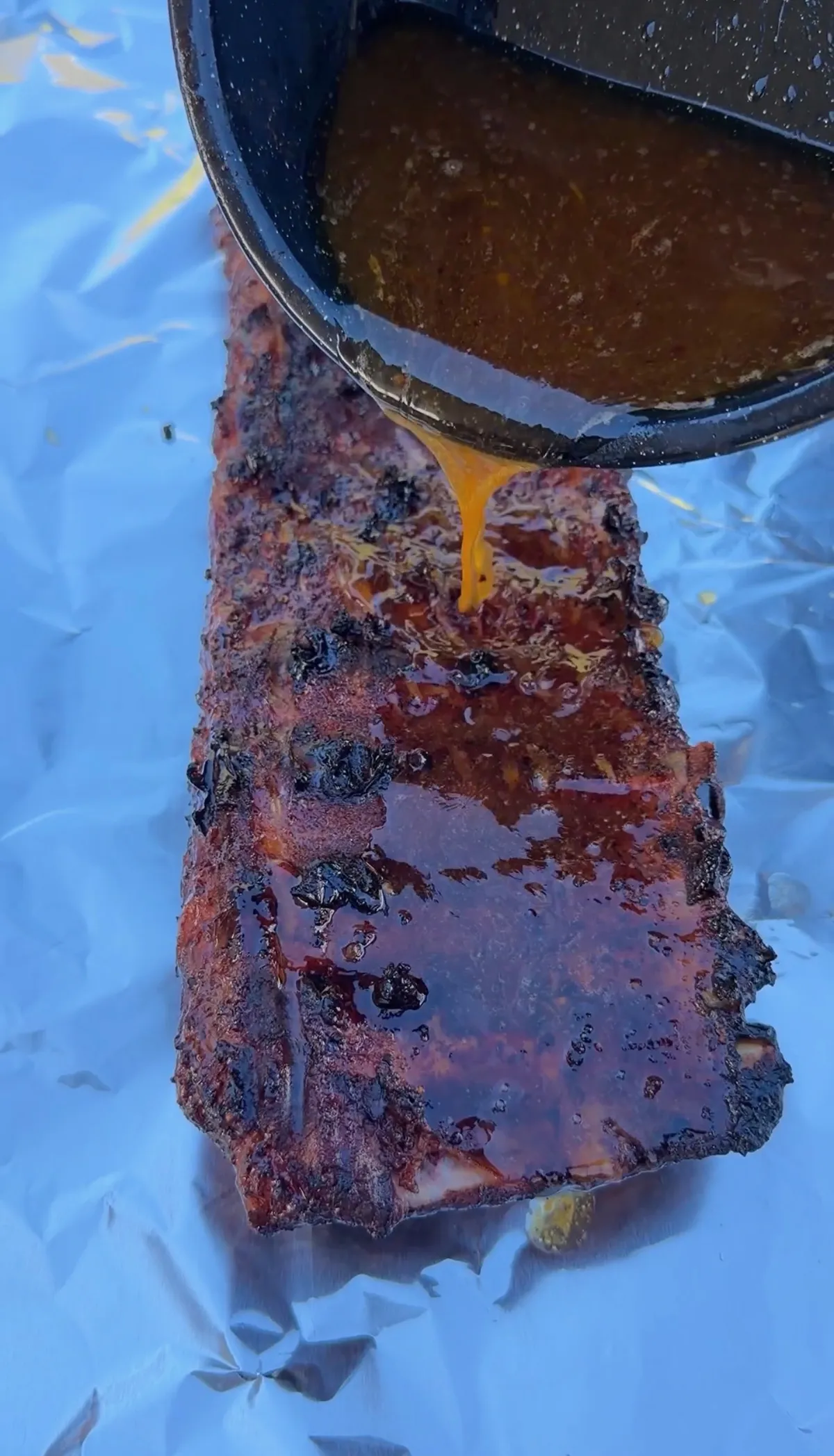 Apple cider BBQ sauce being poured over smoky, caramelized St. Louis spare ribs on aluminum foil