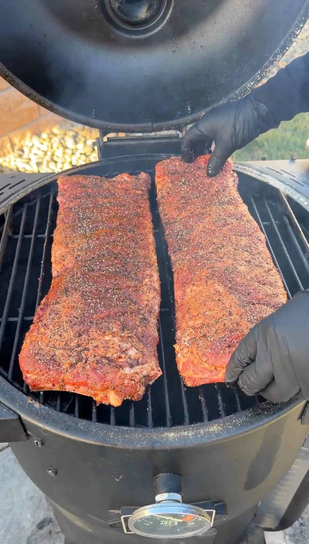 Two racks of dry-rubbed St. Louis spare ribs placed on a charcoal smoker grill, handled with black gloves