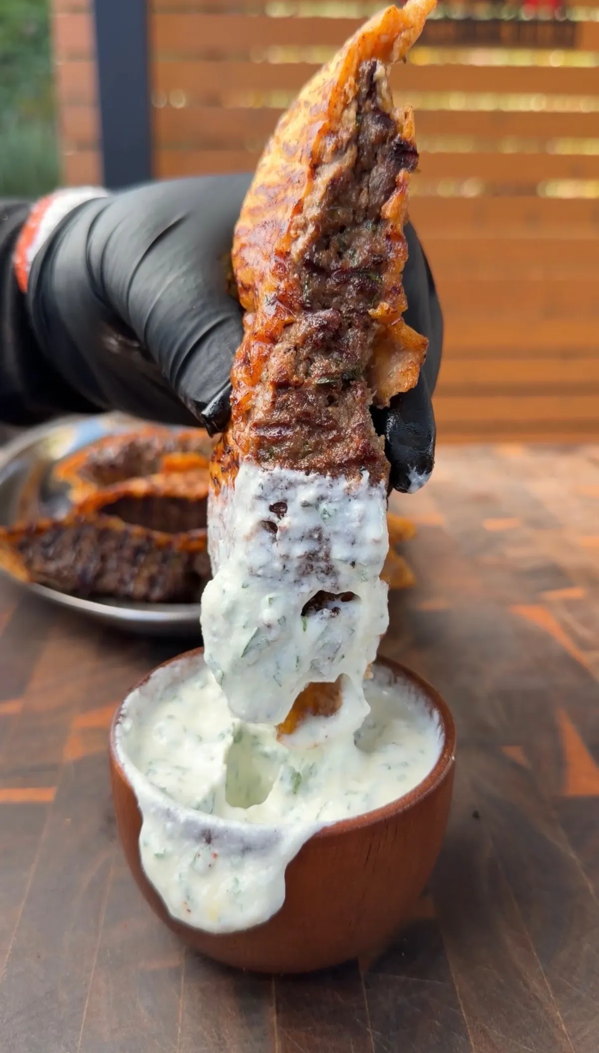 Hand in black glove dipping a crispy arayes pita into garlic yogurt sauce with fresh parsley in a clay bowl