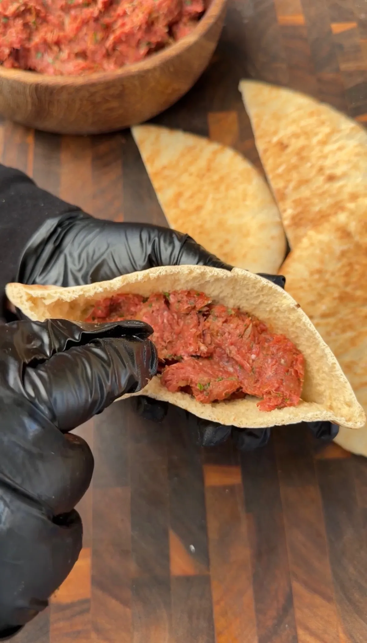 Hands in black gloves opening a pita pocket stuffed with seasoned ground beef mixture for arayes on a wood cutting board