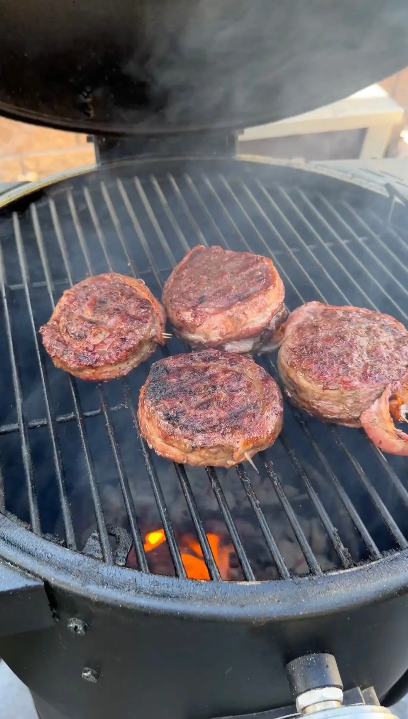 Four bacon-wrapped burger pinwheels grilling over charcoal flames on a round ceramic kamado grill