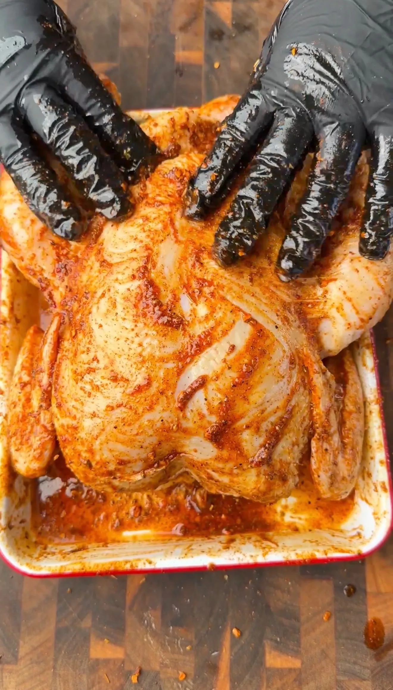 Hands in black gloves rubbing a Cajun oil-and-seasoning mud rub onto a whole raw chicken in a roasting pan
