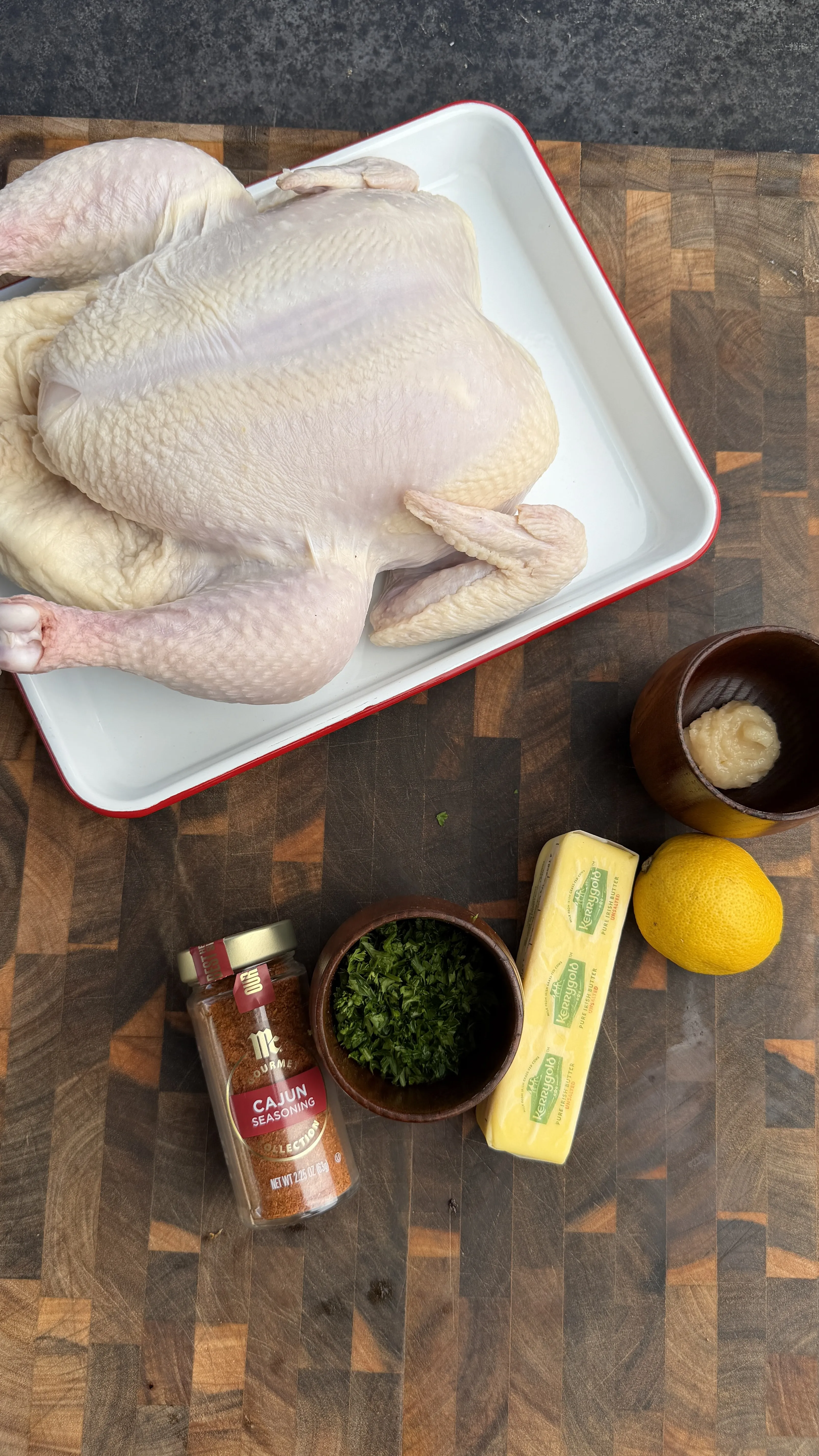 Raw whole chicken on a white tray with Cajun seasoning, butter, parsley, lemon, and garlic on a cutting board
