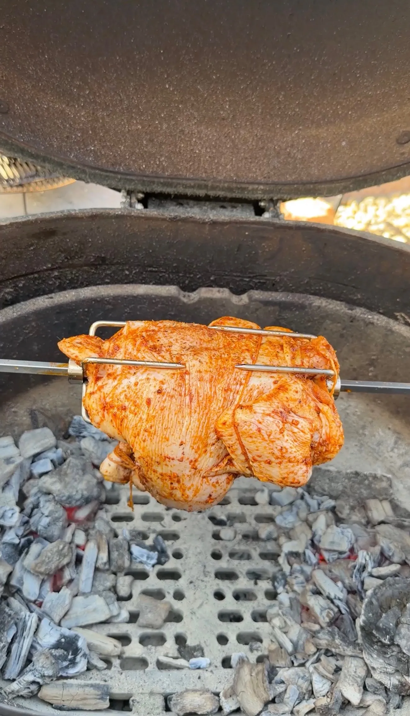 Whole chicken coated in Cajun spice rub mounted on a rotisserie spit over glowing charcoal in a kettle grill