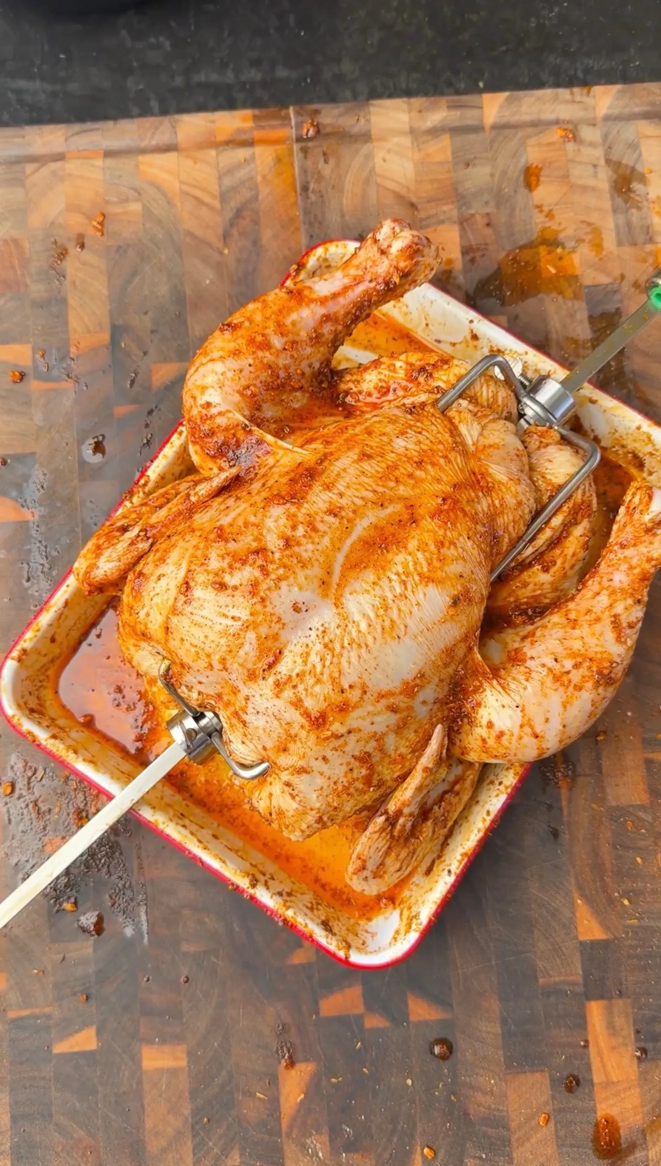Whole Cajun-seasoned chicken on a rotisserie spit in a roasting pan on a wooden cutting board