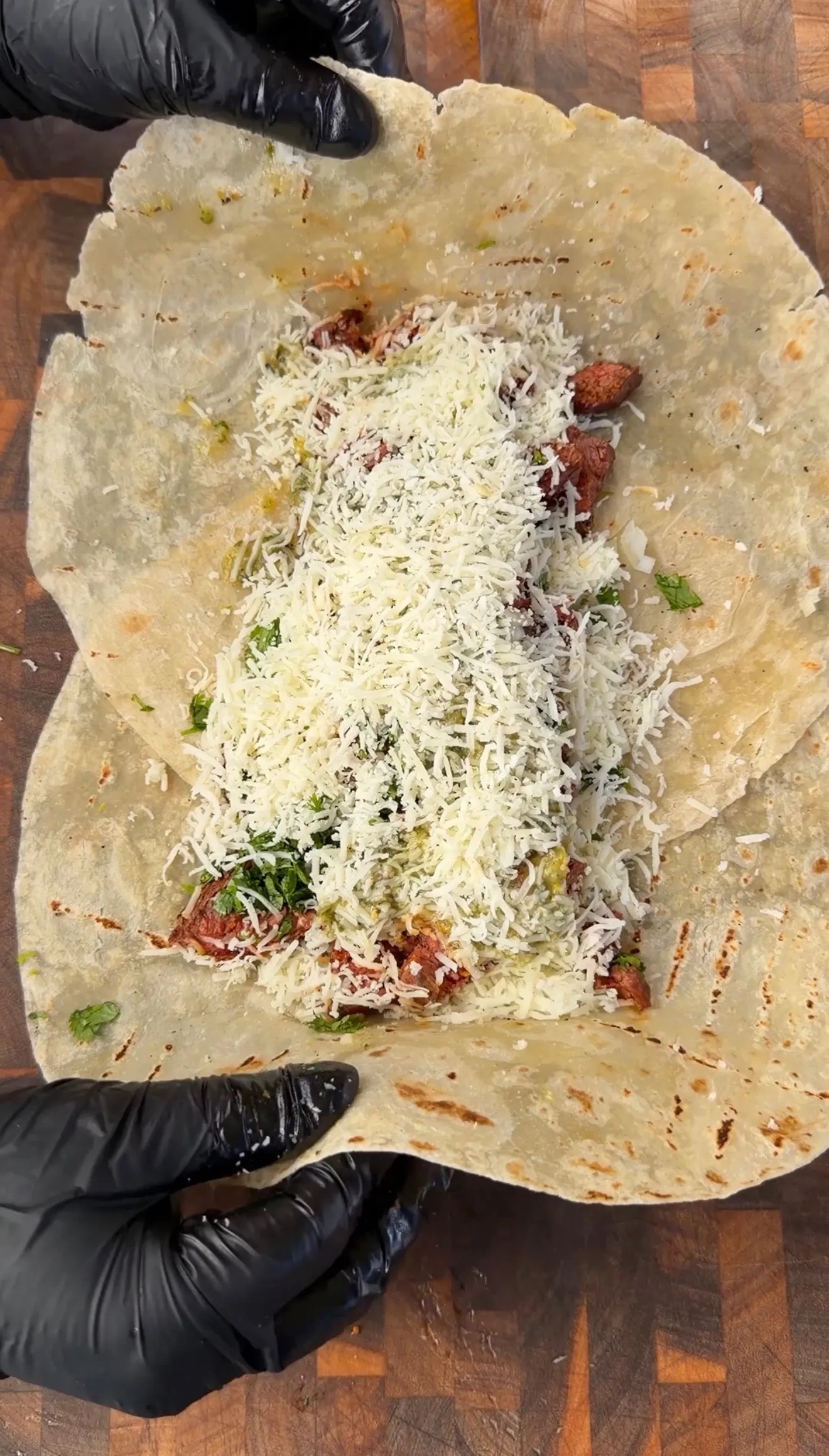 Hands in black gloves wrapping a flour tortilla filled with carne asada, guacamole, cilantro, and shredded cheese