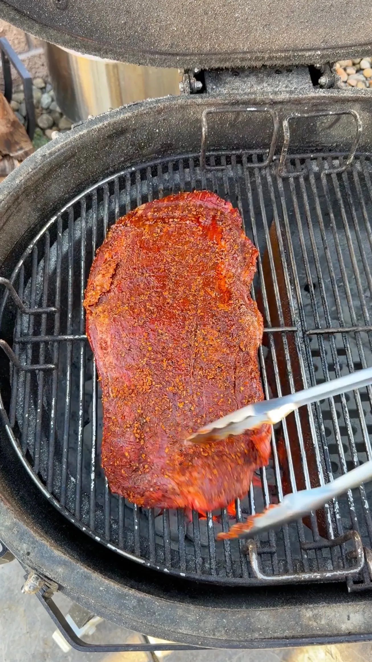 Large beef flank steak coated in red spice rub being grilled on a charcoal kettle grill with tongs