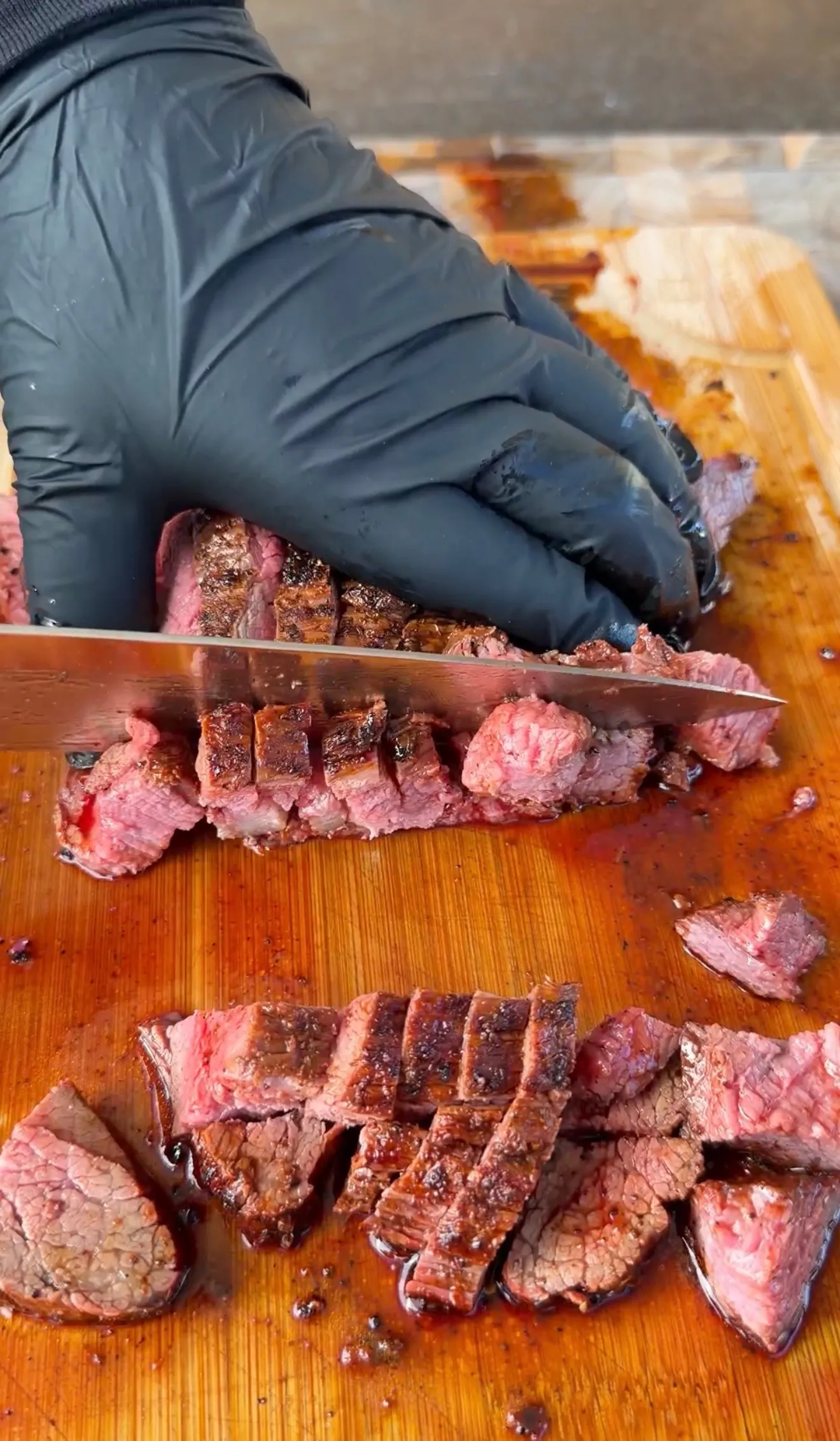 Gloved hands slicing grilled carne asada into chunks on a wooden cutting board with meat juices