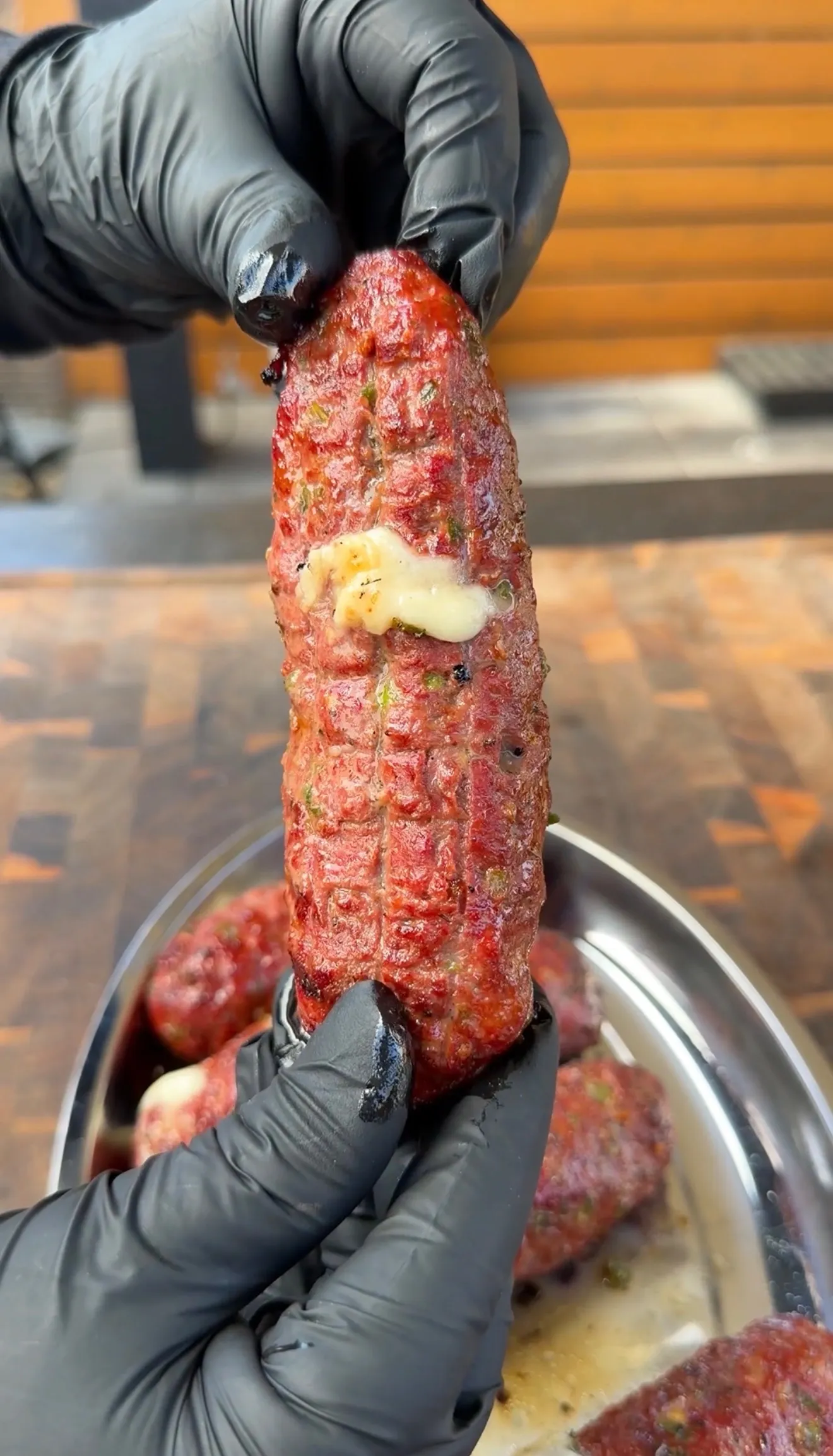 Hands in black gloves holding a grilled cheese-stuffed burger dog with melted cheese oozing out over a metal tray