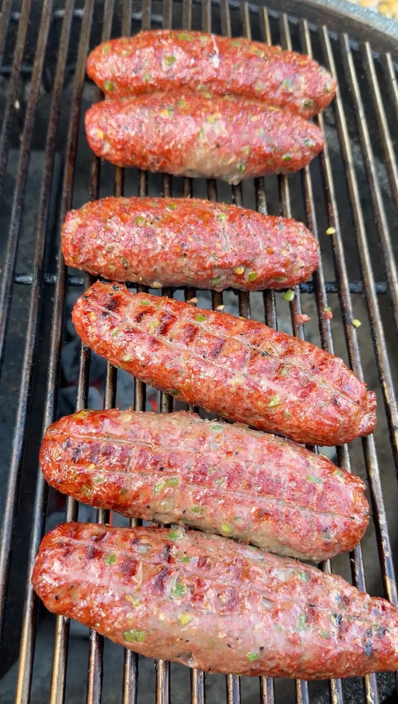 Six cheese stuffed burger dogs cooking on a charcoal grill, showing grill marks and herb-speckled meat